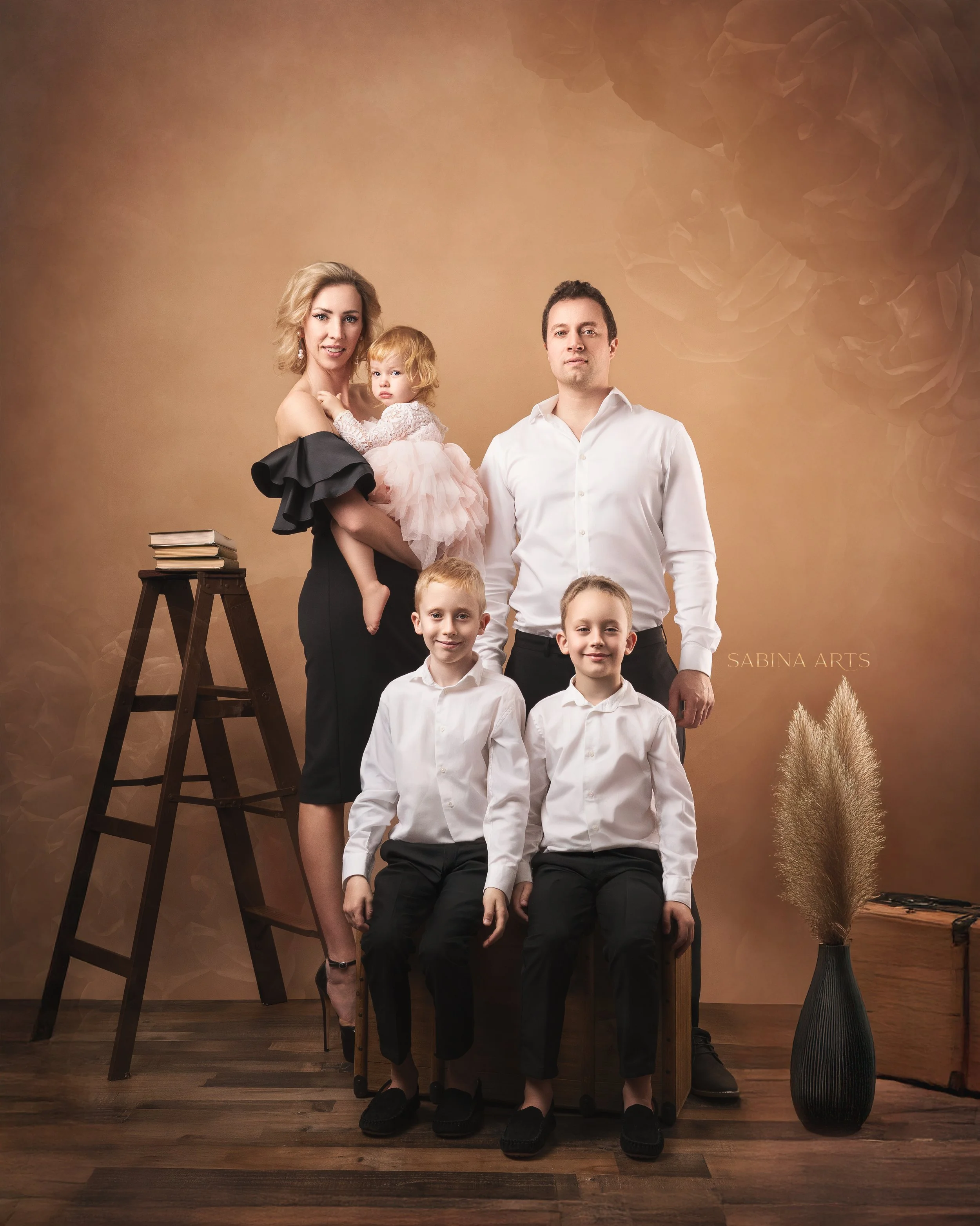 A family of six posing indoors with a warm brown background. The mother is holding a young girl in a pink dress, and four boys in white shirts are in front. There is a black ladder with books on it on the left and a tall dark vase with beige dried pl