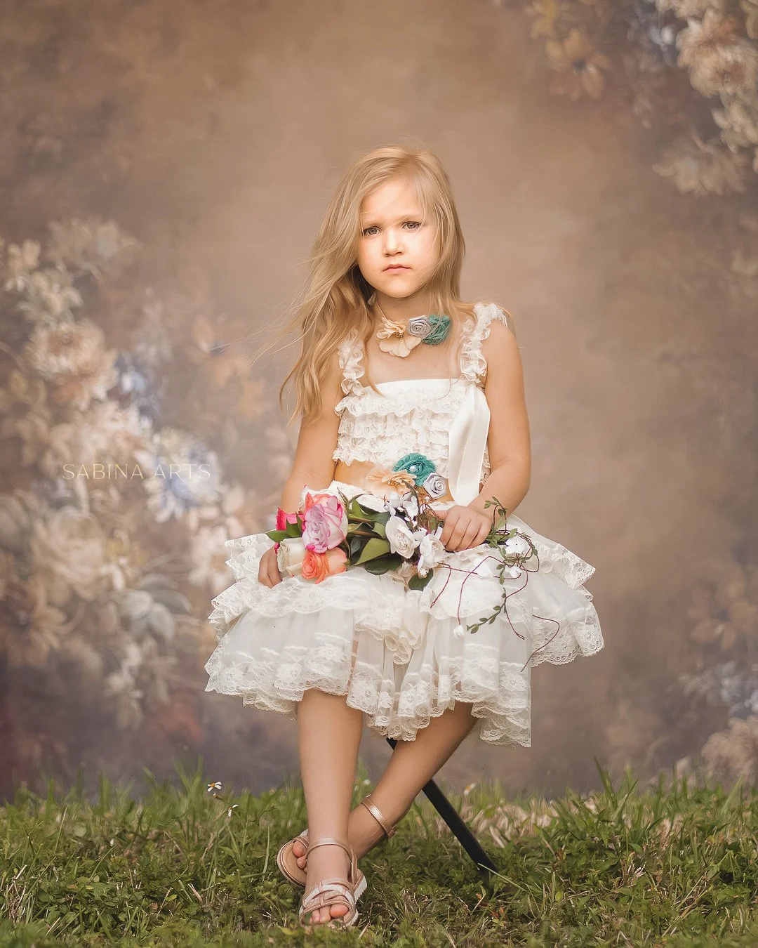 A young girl with long, blonde hair sitting outdoors on a black stool. She is wearing a white lace dress with ruffles and floral accents, and beige sandals. She holds a bouquet of colorful roses and greenery. The background is a blurred, floral-themed backdrop.