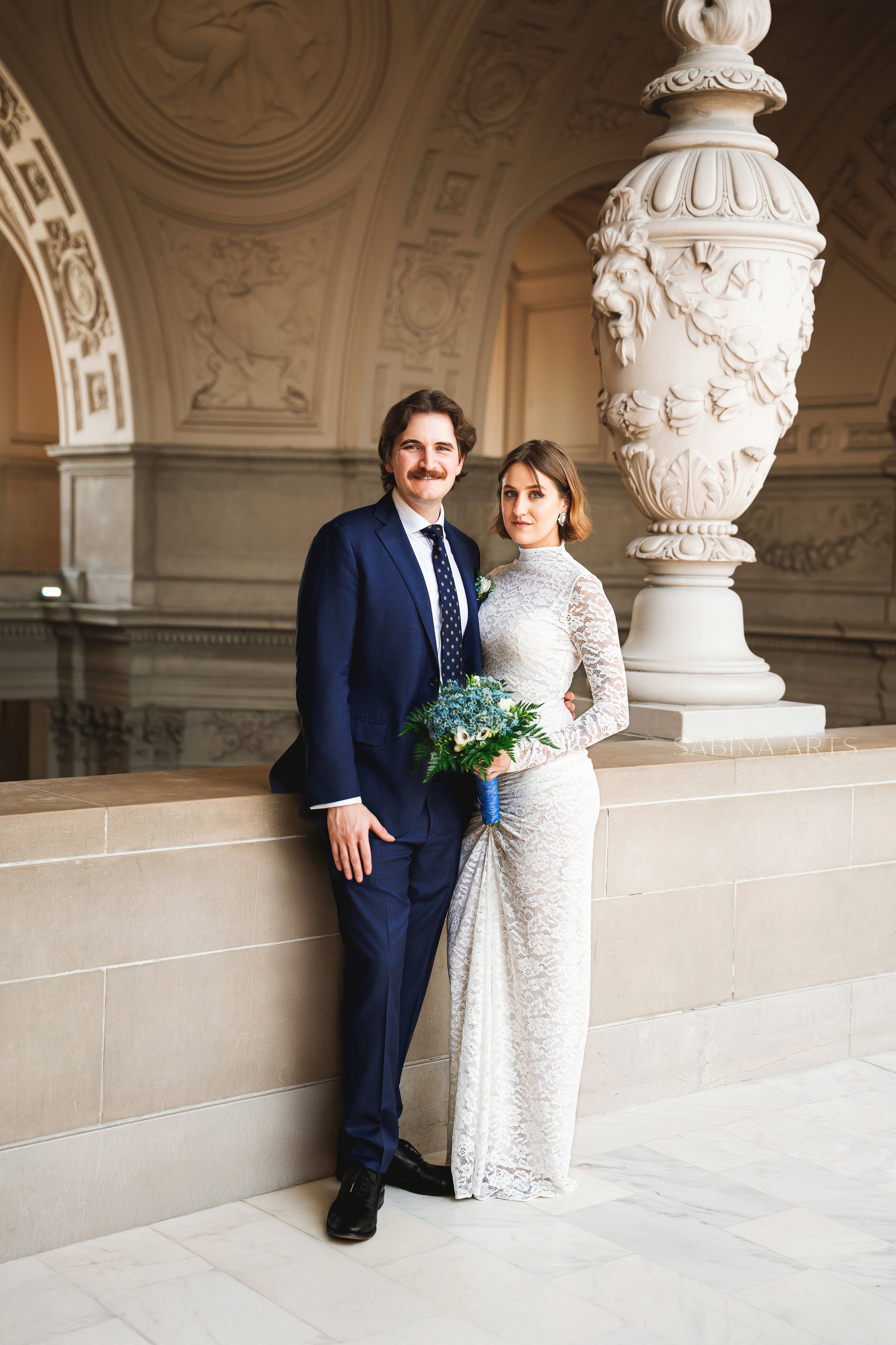 A bride and groom stand close together indoors, with the groom in a navy suit and the bride in a lace white wedding dress holding a bouquet of blue and white flowers. Behind them is a large ornate white vase with a lion's head decoration. The backgro