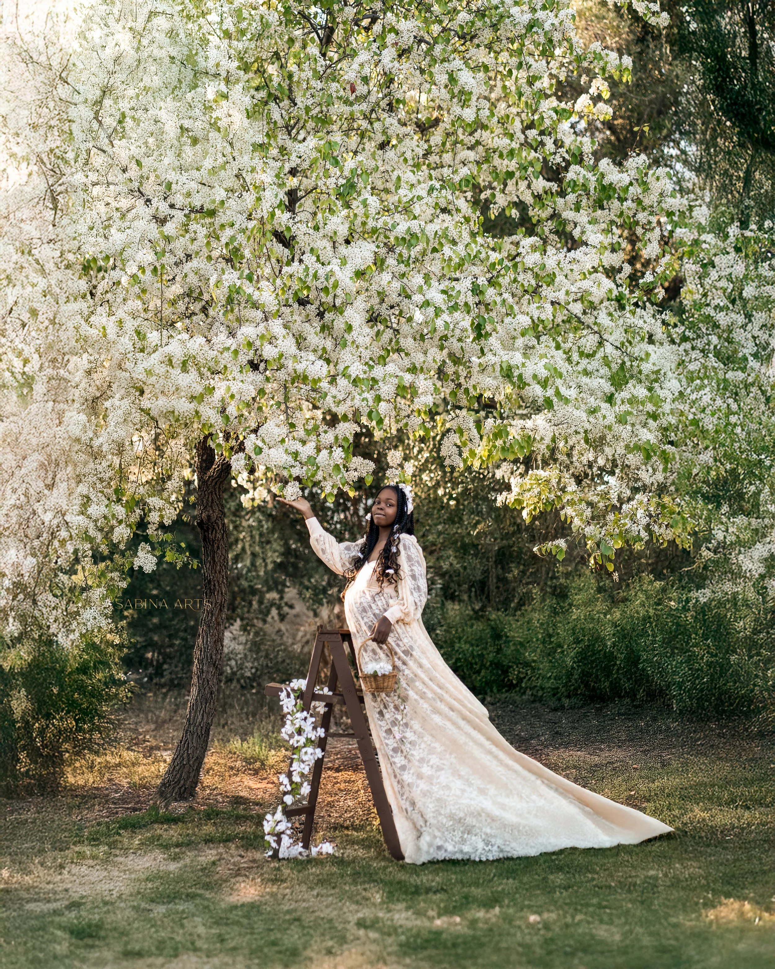 A woman in a long, flowing cream-colored lace dress is standing on a small wooden step ladder under a flowering tree with white blossoms, holding a basket of flowers and reaching out to touch the blossoms. The background shows a lush garden with gree