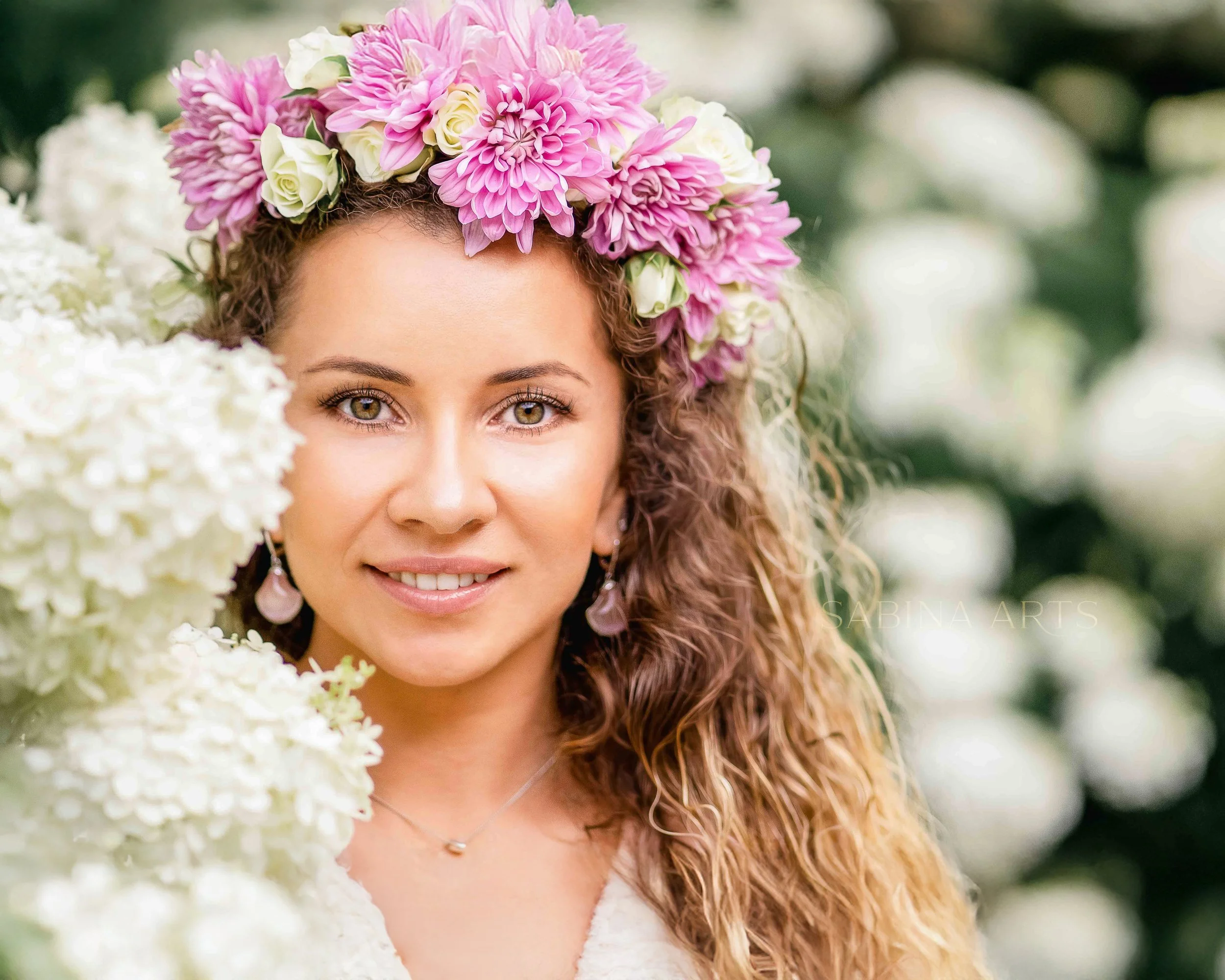 Close-up of a woman with curly hair wearing a floral crown made of pink and white flowers, standing outdoors with a blurred green background.