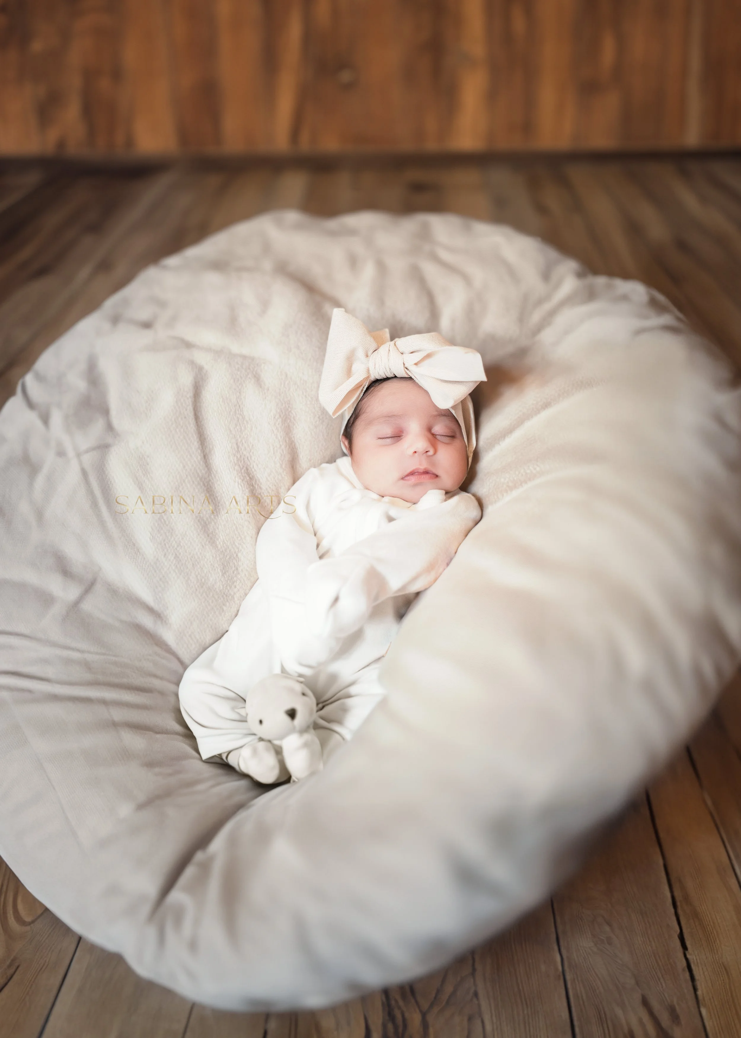 A sleeping baby with a cream-colored headband with a bow, lying on a cream-colored blanket in a round bed, with a small teddy bear nearby, on a wooden floor.