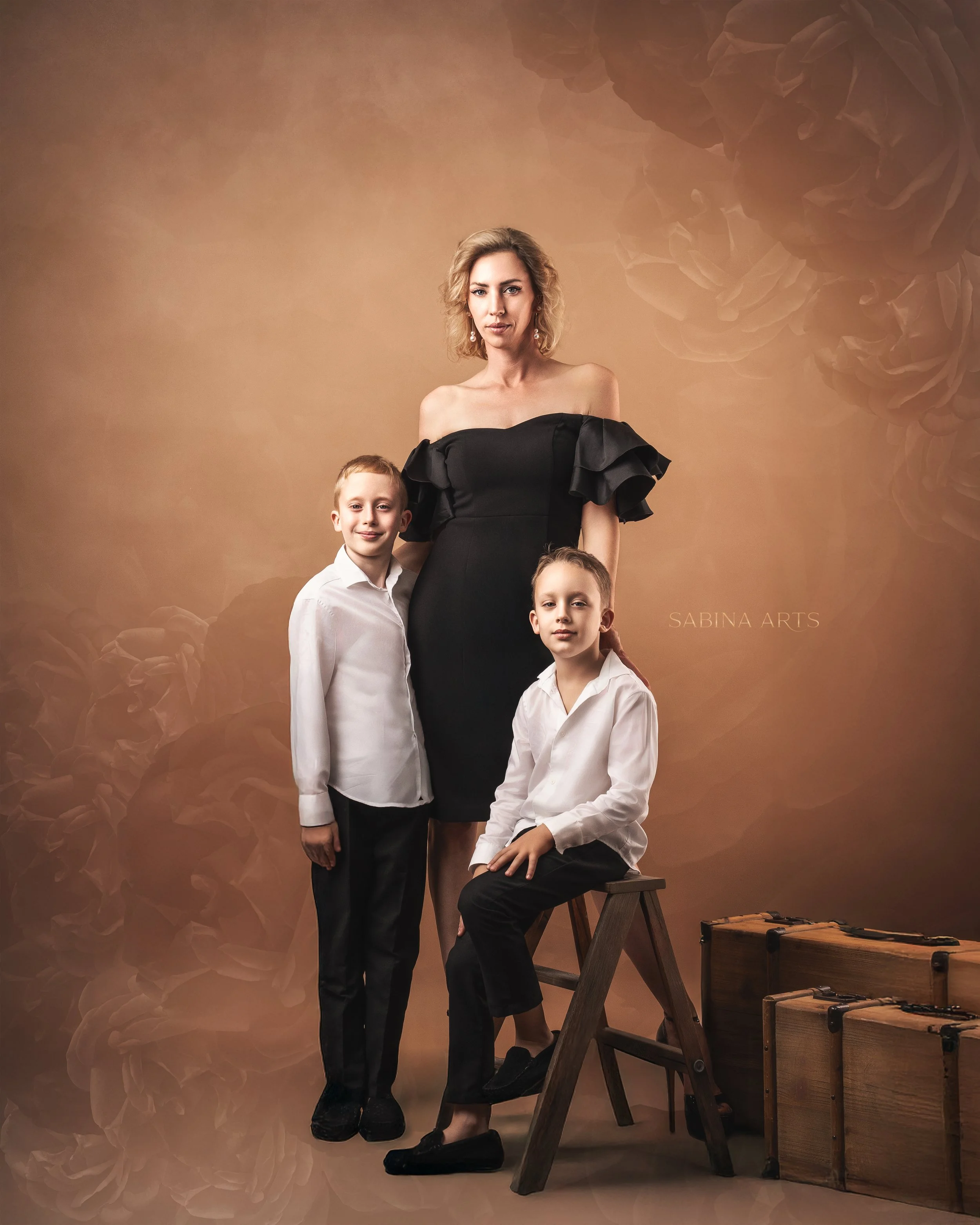 A woman and three young boys posing for a portrait in front of a brown background with floral designs. The woman is standing, wearing a black off-shoulder dress with ruffled sleeves, and has shoulder-length wavy blonde hair. One boy is sitting on a s