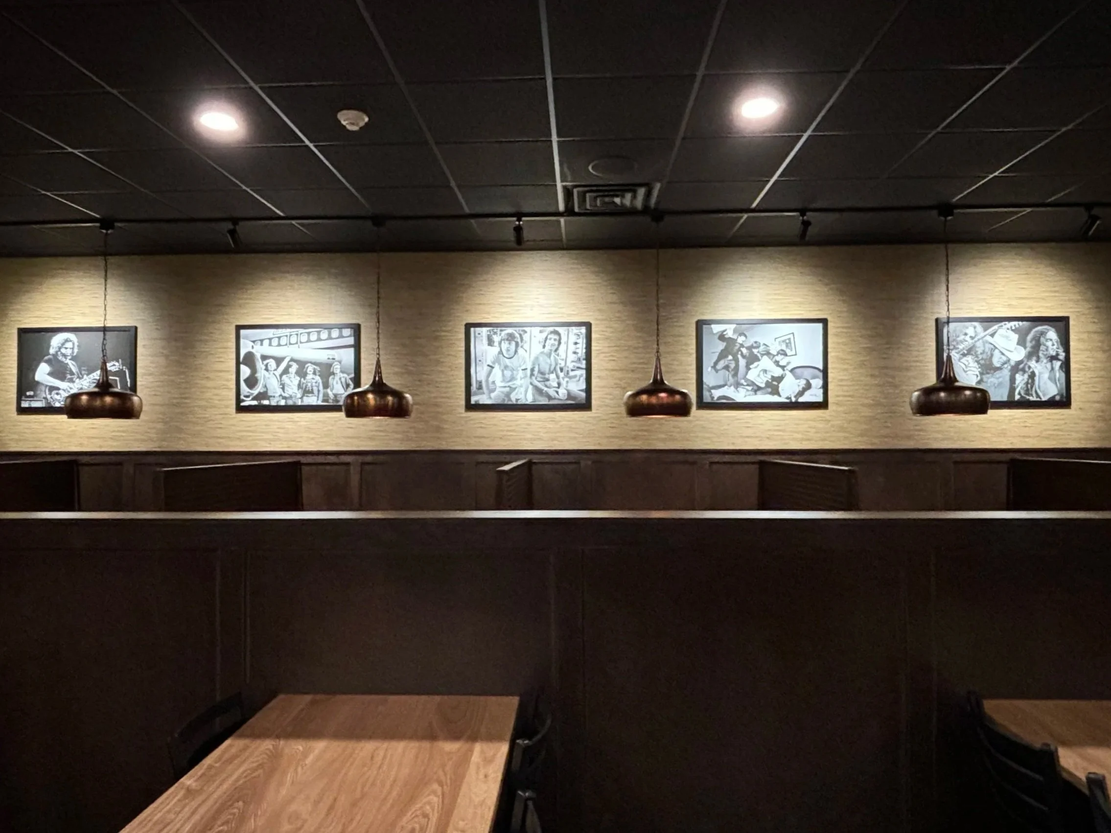 A dimly lit room with a black ceiling and beige walls, decorated with black and white photographs framed on the wall. There are three hanging pendant lights with a copper finish illuminating the space. Empty chairs are arranged in the room, and there is a wooden table in the foreground.