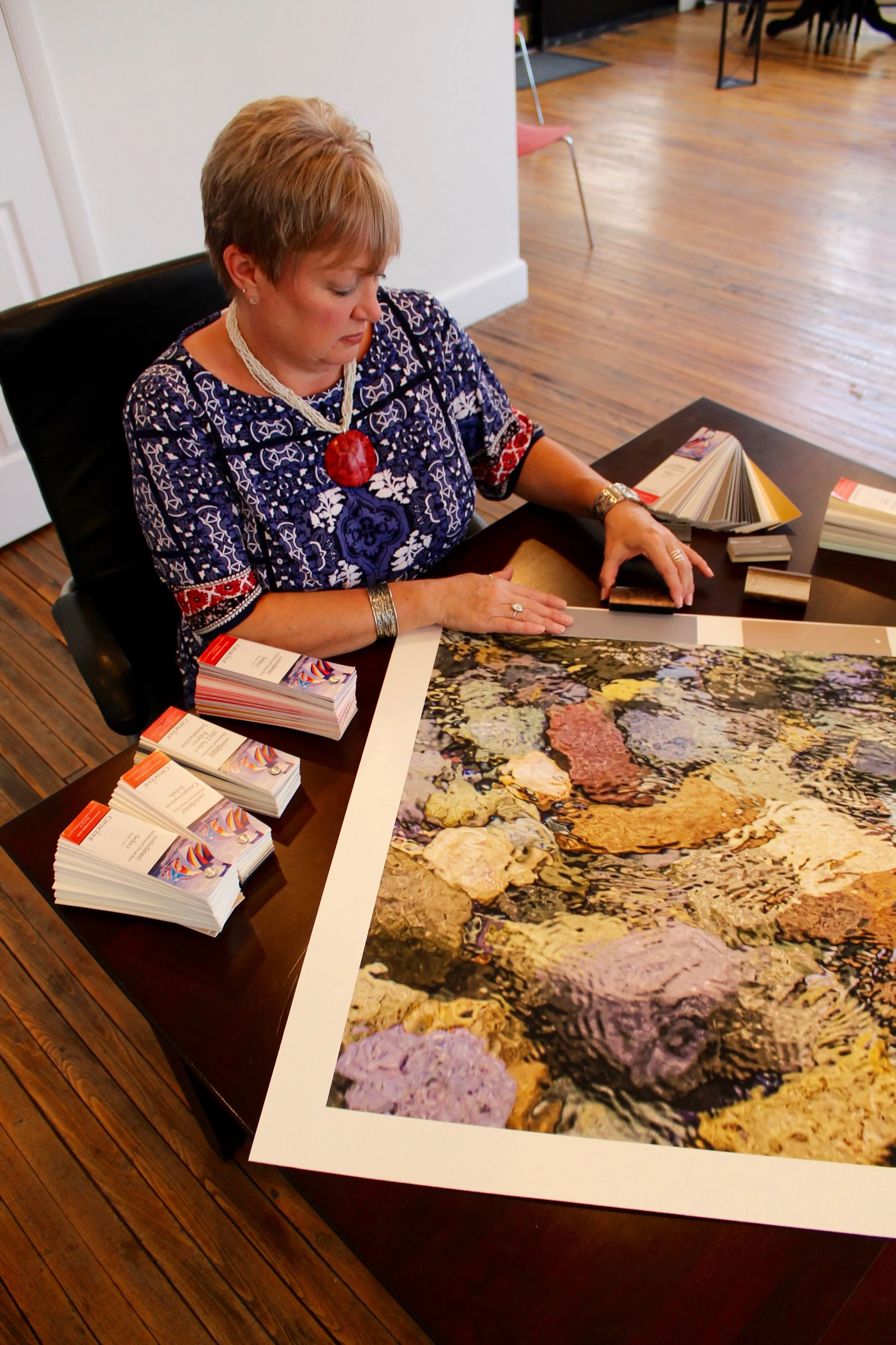 A woman with short hair wearing a blue and white patterned dress, a large red necklace, and silver jewelry, sits at a table looking at color samples and fabric swatches. A table in front of her displays brochures with colorful graphics and a large print of a flower garden with various flowers and rocks.