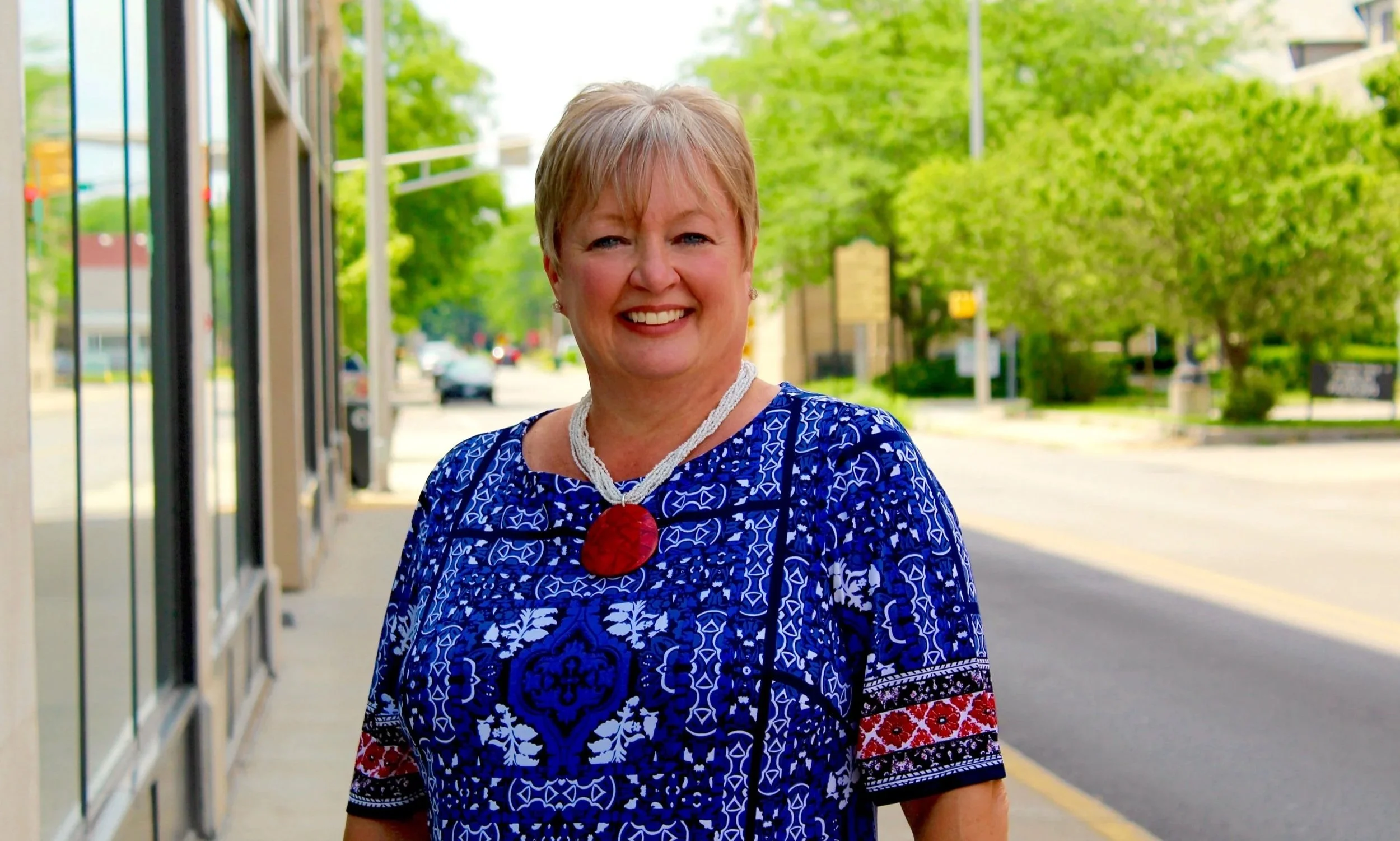 A smiling middle-aged woman with short blonde hair, wearing a vibrant blue patterned dress and a large red necklace, stands outdoors on a sidewalk near a storefront, with green trees and buildings in the background.