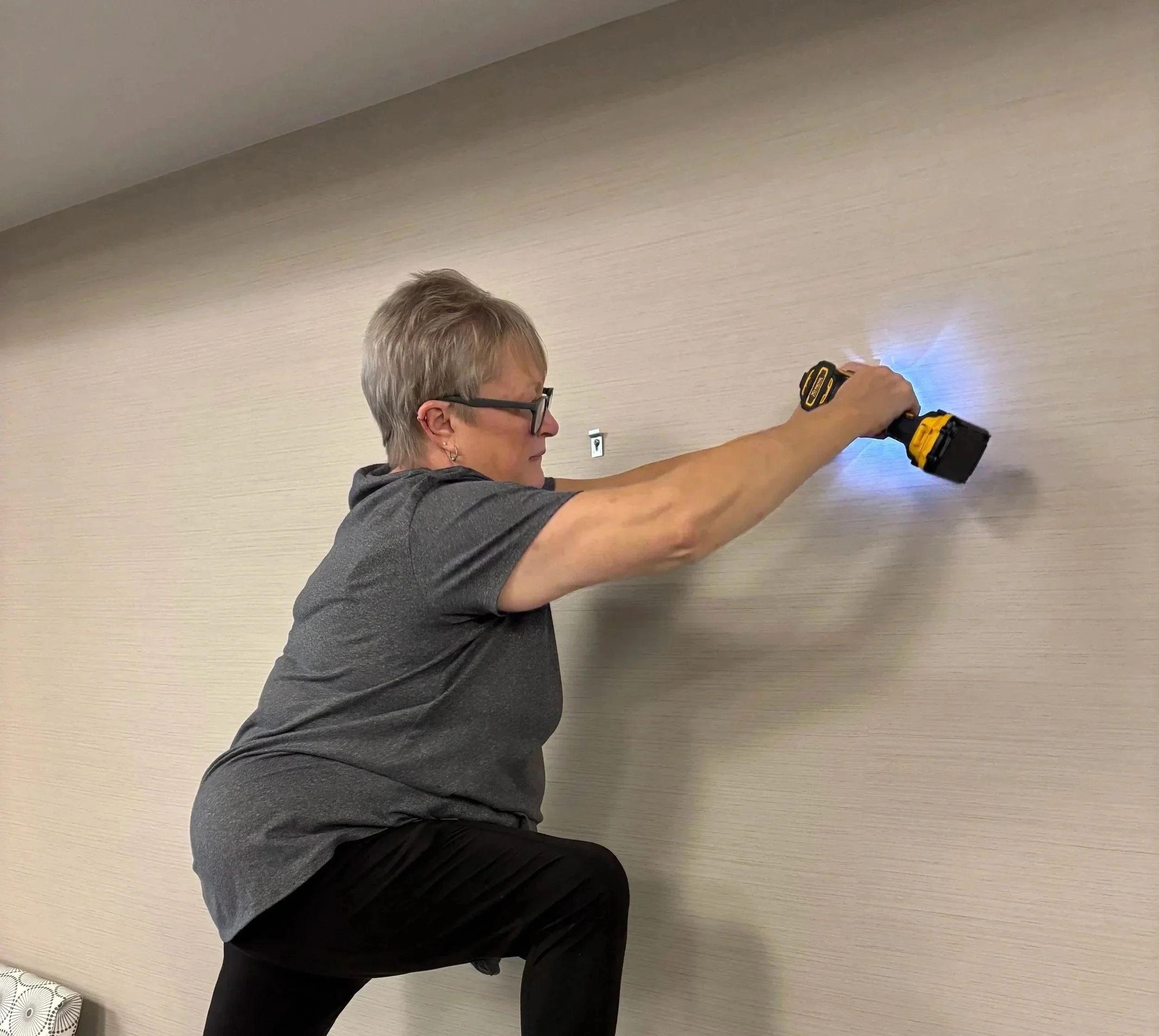 A woman with short gray hair and glasses, wearing a gray shirt, is using a yellow cordless drill on a beige wall in a room with a white electrical outlet and a wall decoration in the corner.