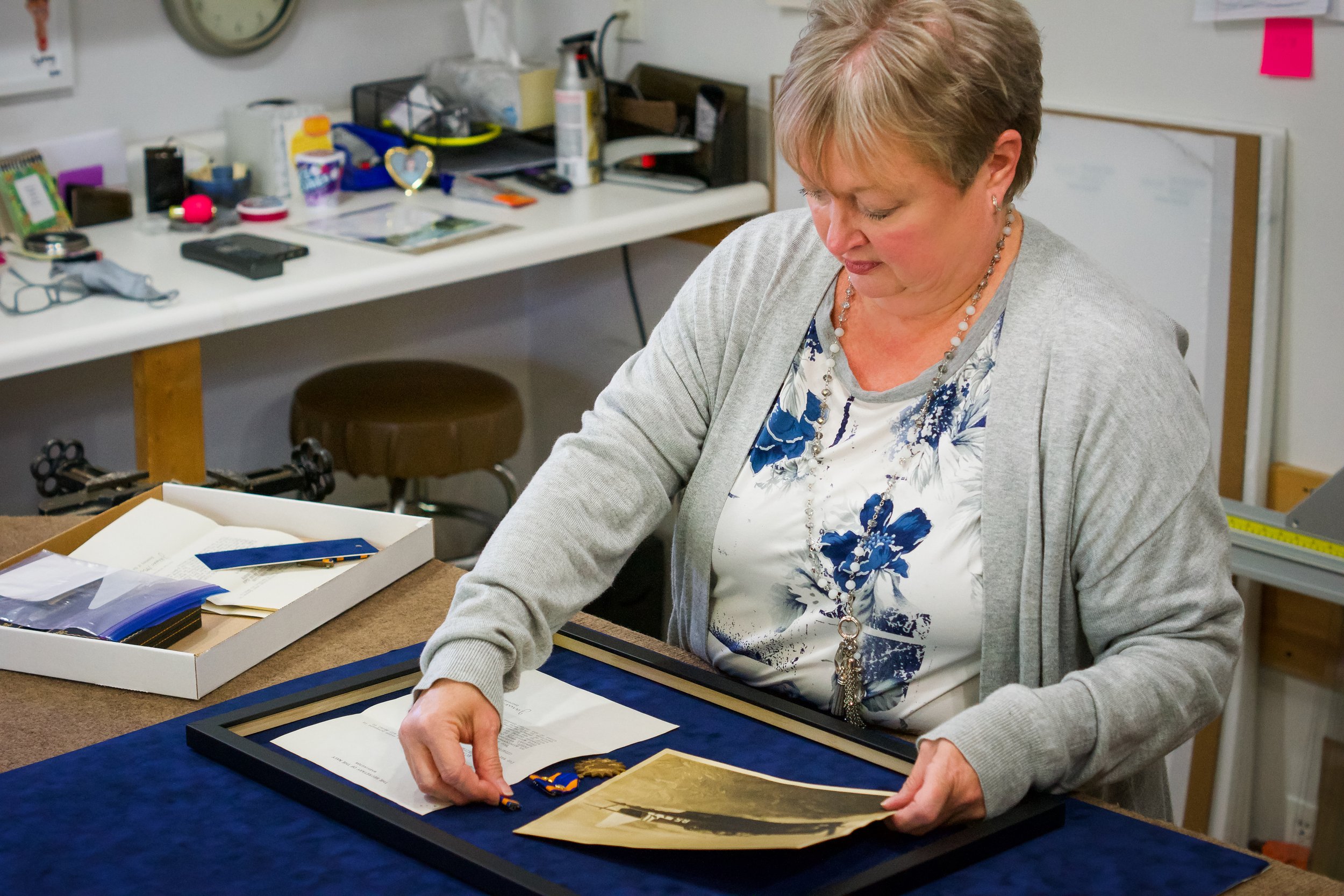 A woman with short blonde hair, wearing a grey sweater over a floral blouse and a necklace, is looking down while arranging framed photos and awards on a table in an office or craft room. Behind her, there is a cluttered white desk with various office supplies, a clock, and a pink sticky note on the wall.