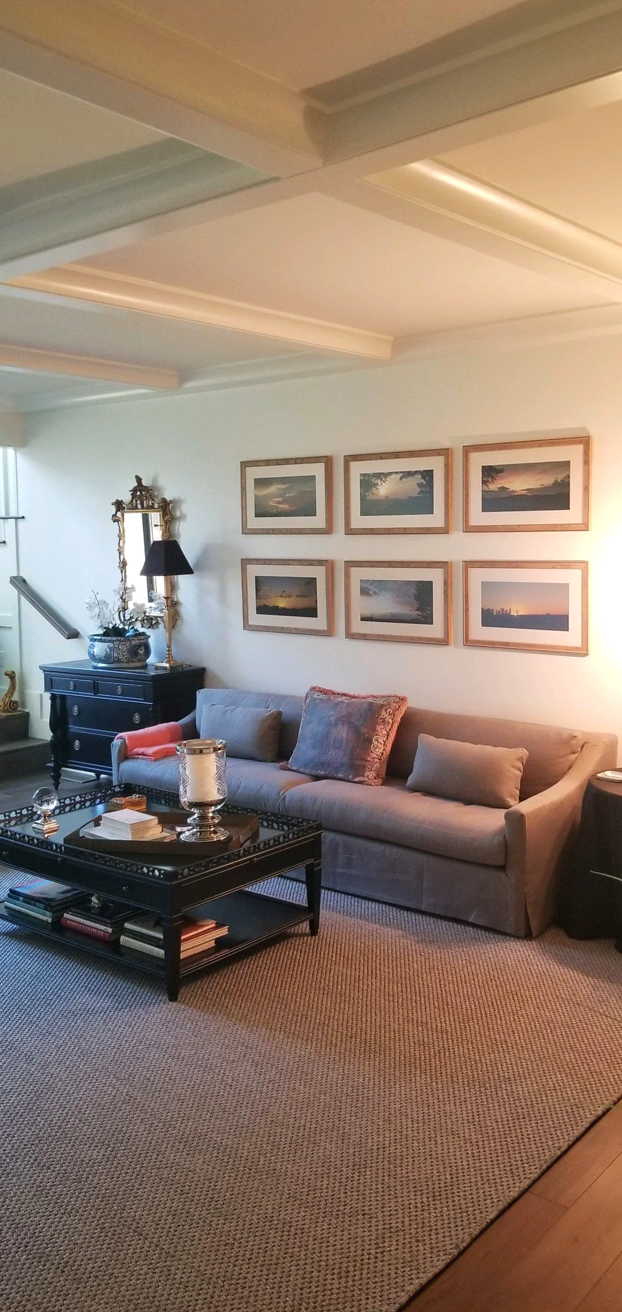 Living room with a gray sofa, framed photographs of sunsets and landscapes on the wall, a black coffee table with books and decorative items, a black side cabinet with a blue vase and a mirror, and a beige carpet.