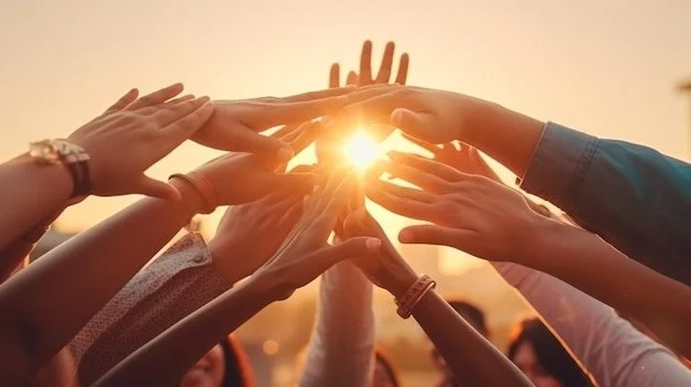 Multiple hands reaching towards the sunlight during sunset, forming a heart shape.
