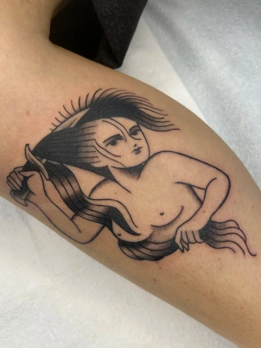 Black ink tattoo of a stylized mermaid with long hair, holding a comb in one hand and resting her other hand on her hip.