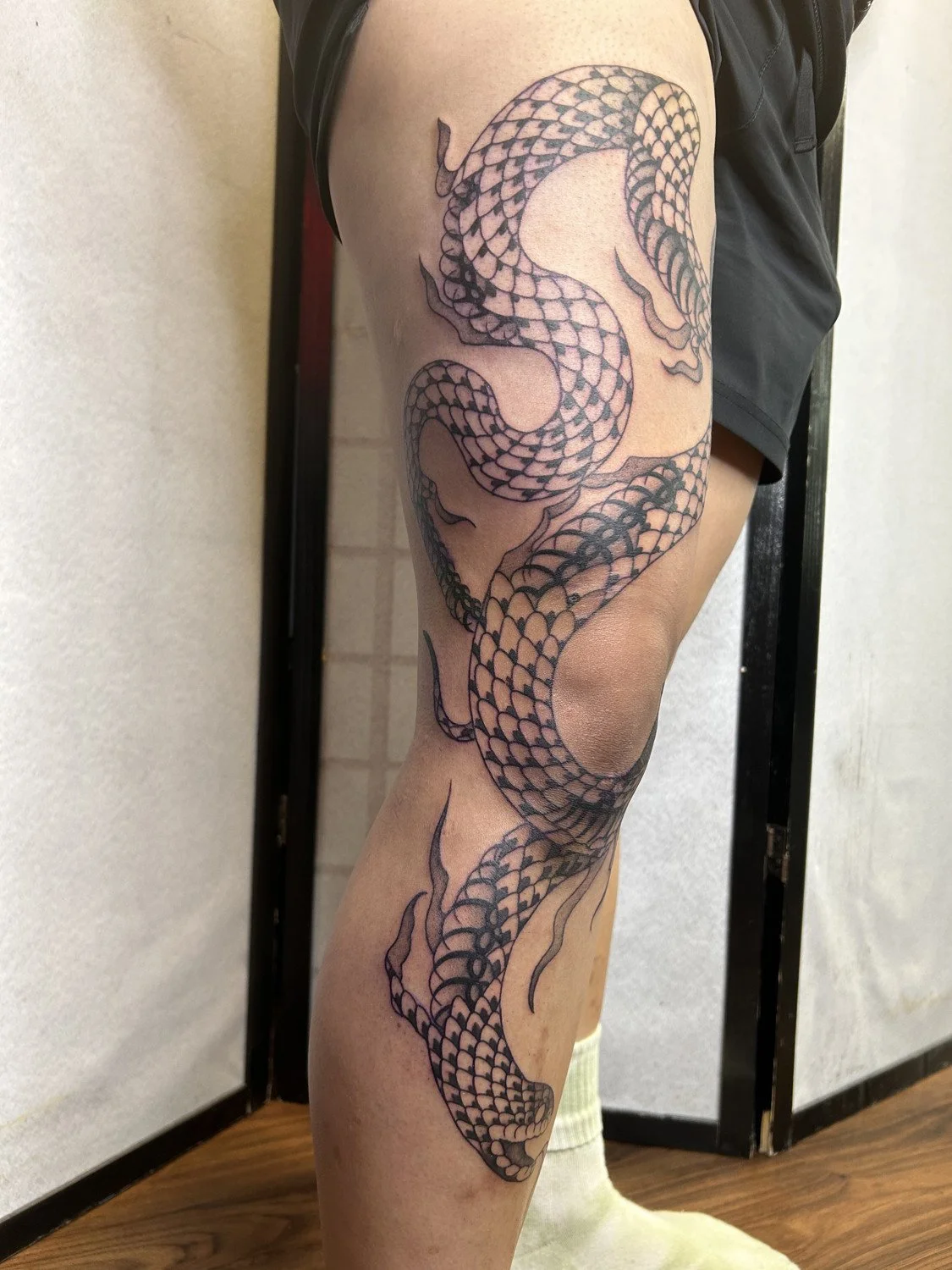 A person with a detailed black ink snake tattoo on their thigh and calf, depicting a coiled snake with scales.