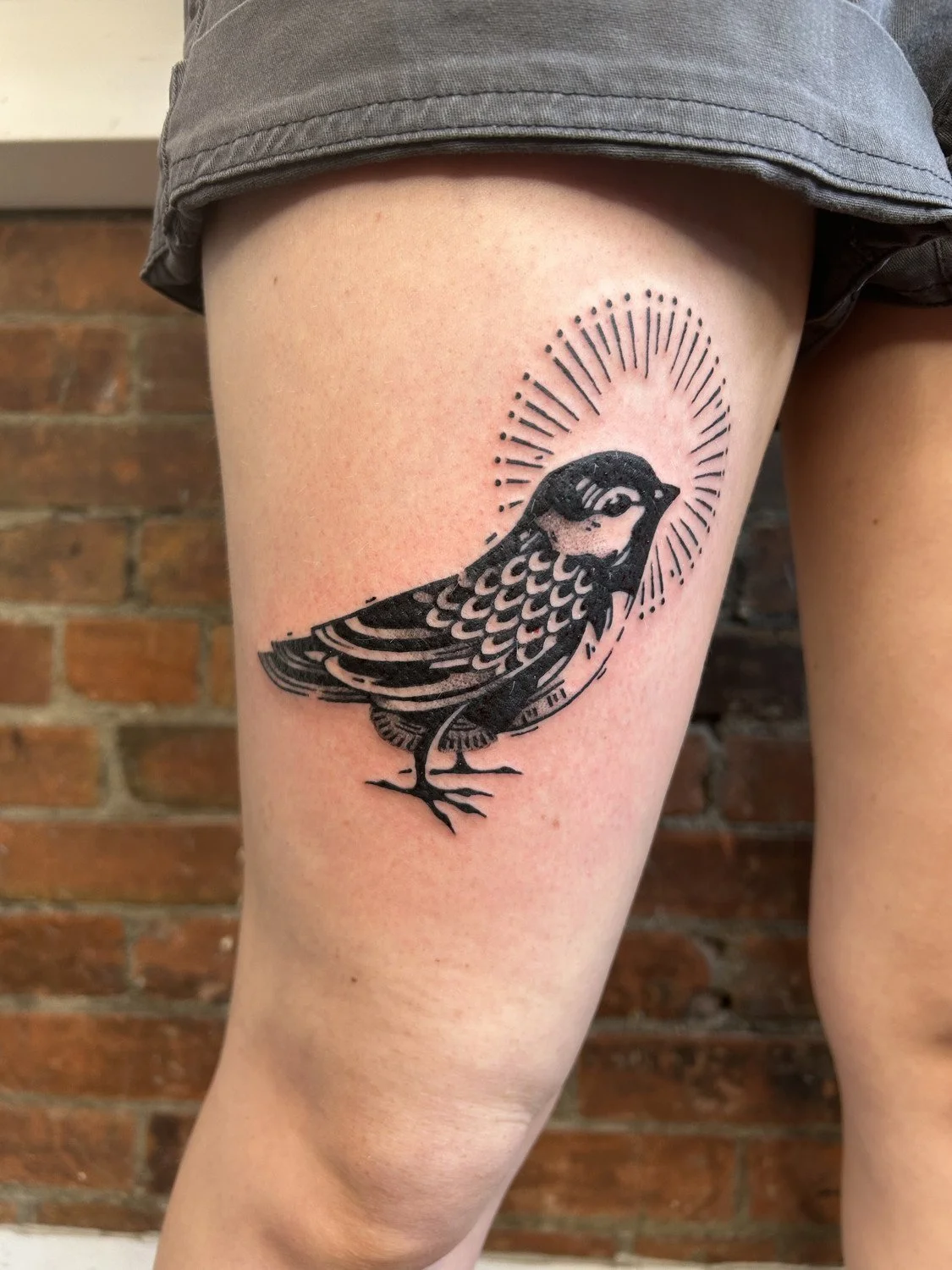 Tattoo of a bird with a halo of radiating lines around its head on a person's thigh.