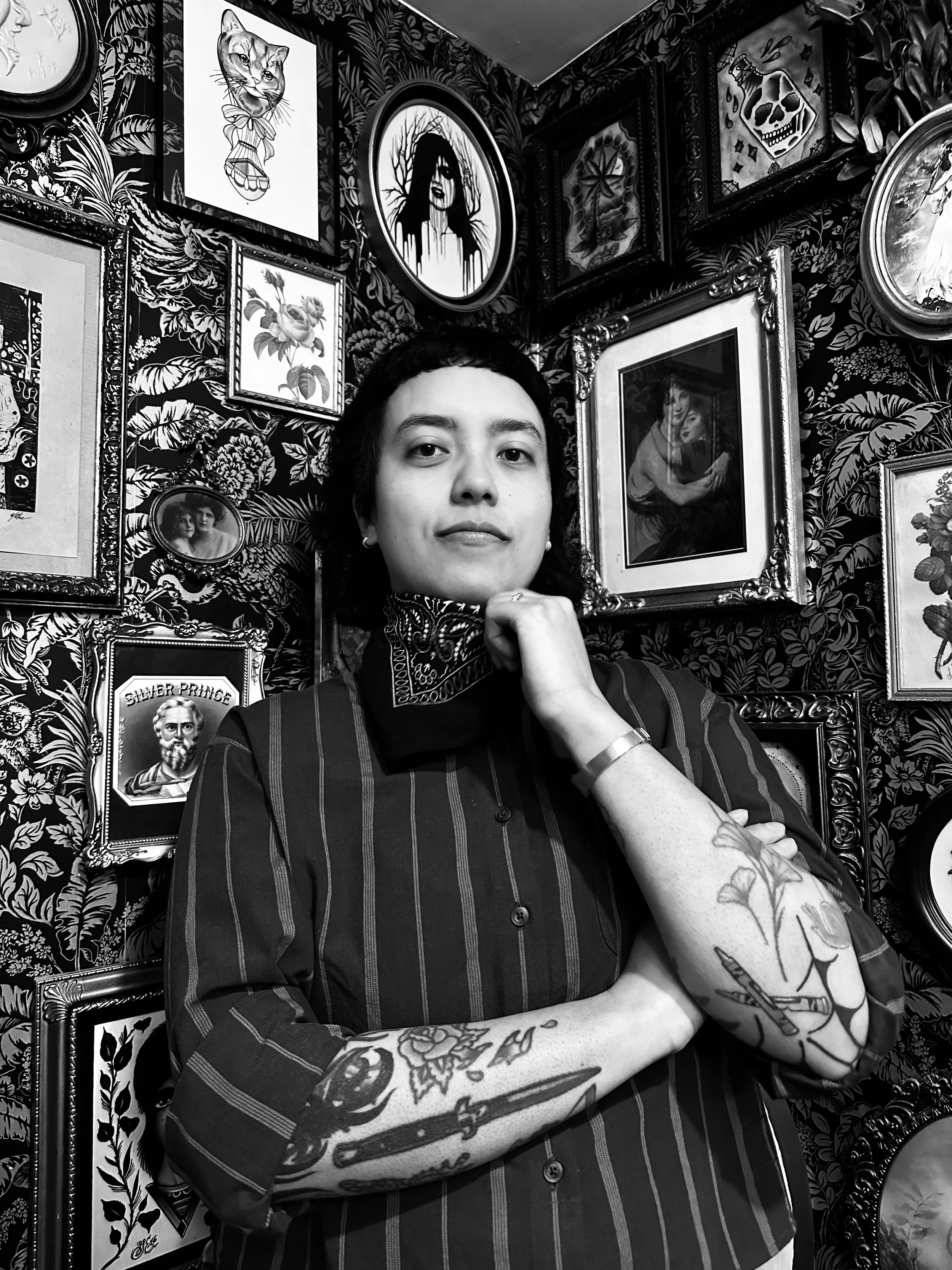 A woman with short dark hair, wearing a striped shirt, tattoos on her left arm, and a scarf around her neck, standing in front of a wall decorated with framed artwork and photographs, in black and white photography.