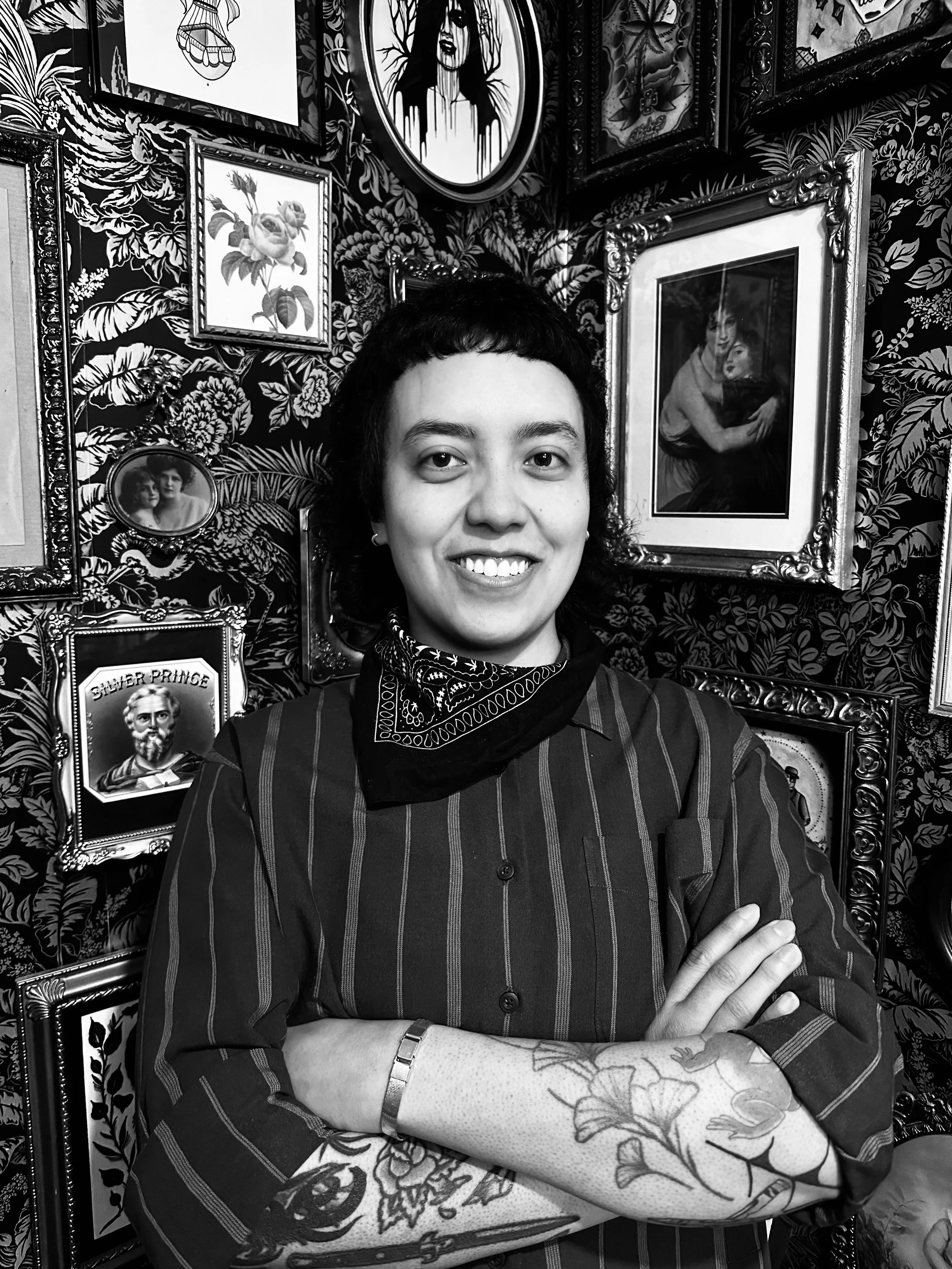 A person with tattoos on their arms, wearing a striped shirt, crossing their arms with a smile, standing in front of a wall decorated with various framed pictures and artwork, in black and white.