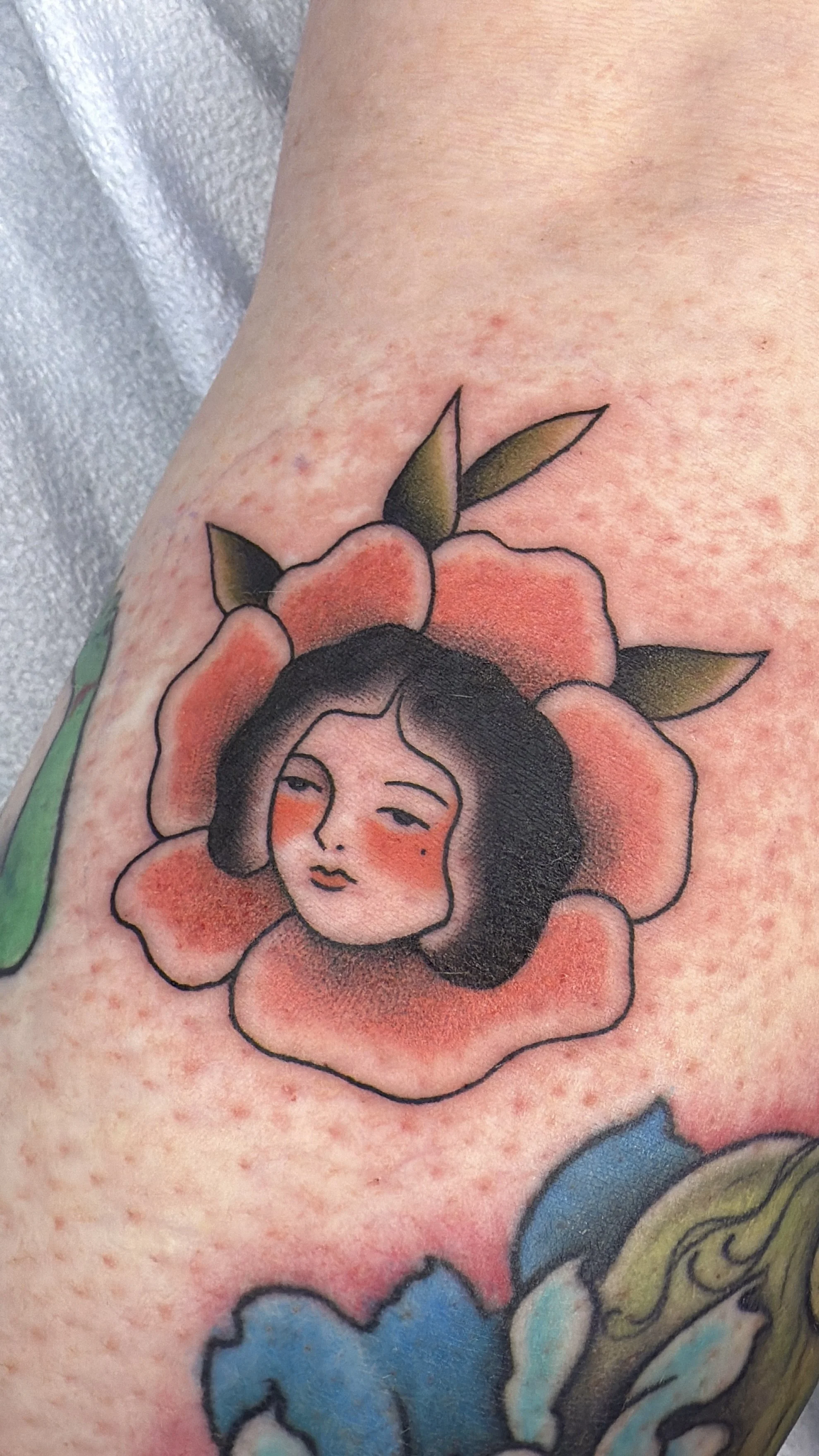 Close-up of a tattoo of a woman's face with dark hair, surrounded by orange and pink flower petals and green leaves.