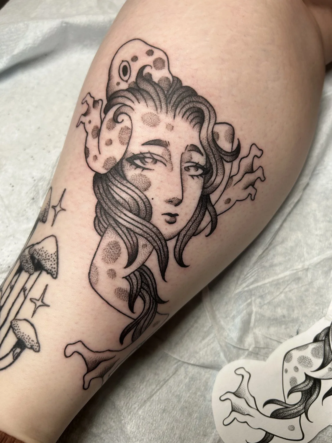 Tattoo of a woman's face with flowing hair, surrounded by mushrooms and abstract elements.
