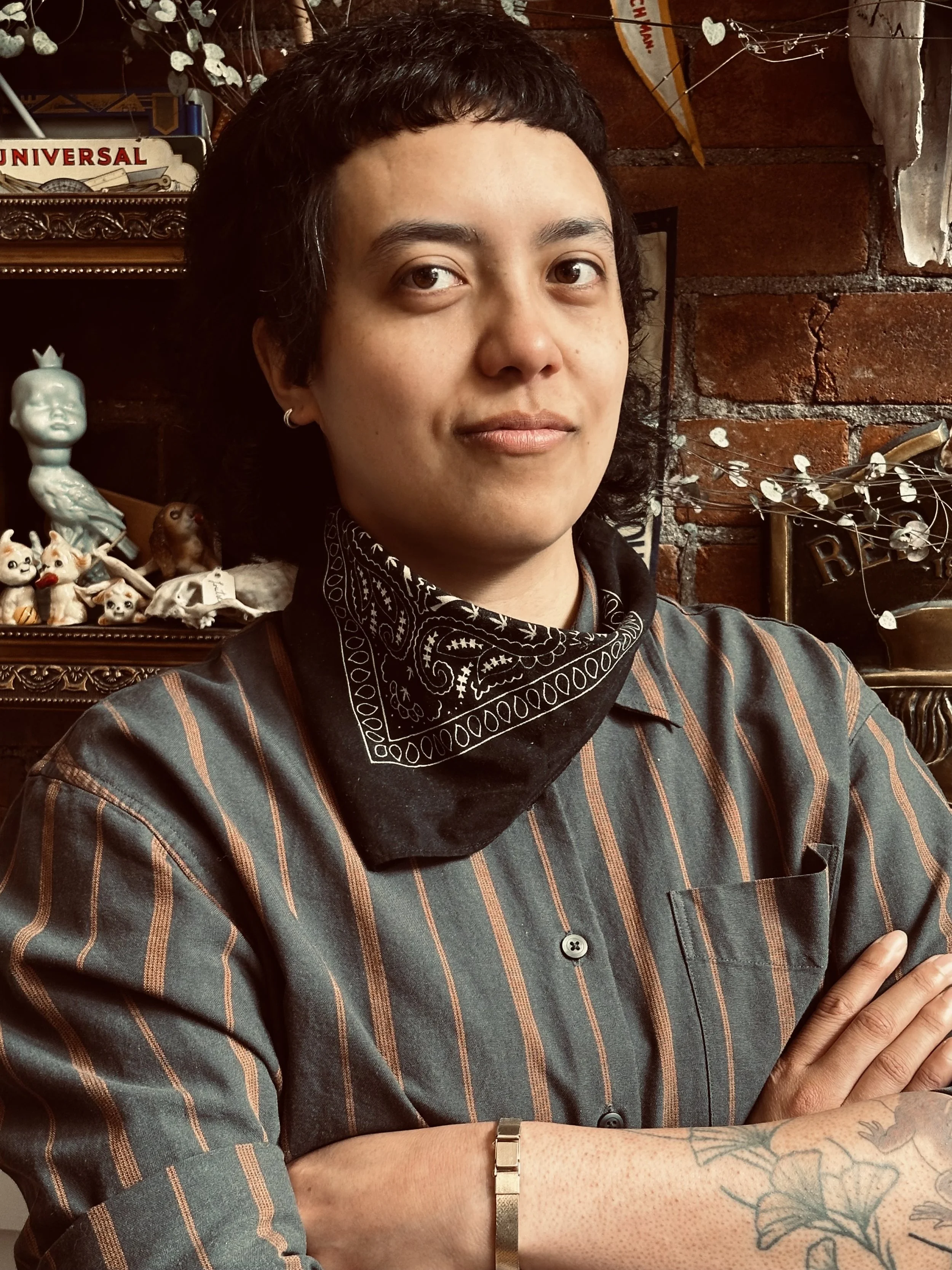 A person with short black hair and light skin, wearing a striped shirt and bandana, standing in front of a brick wall with decorative items and signs, including a small statue and figurines.