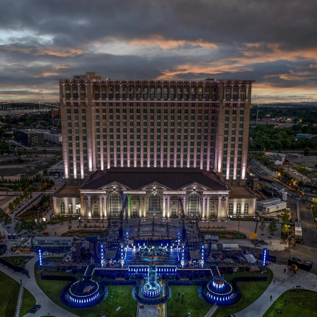 A large hotel building with vertical white lights and a decorated stage in front, under a cloudy sunset sky.