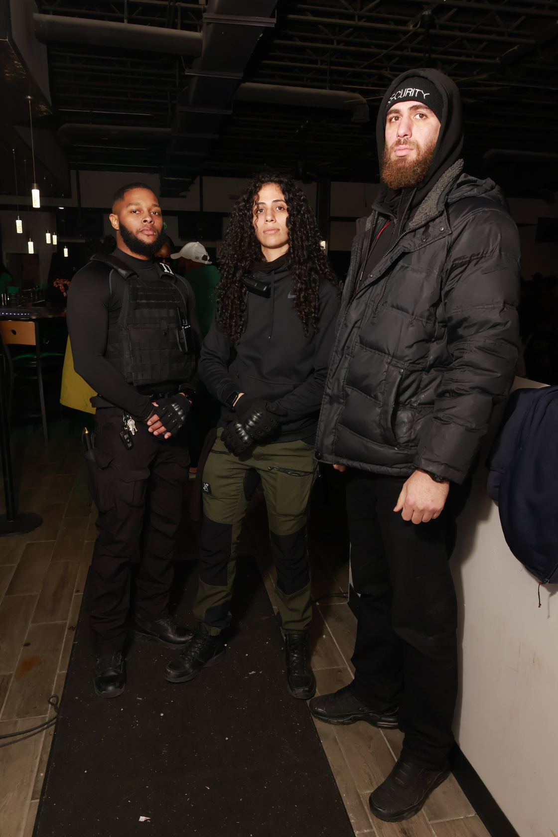 Three security personnel standing inside a building, dressed in black tactical and winter gear, with serious expressions.