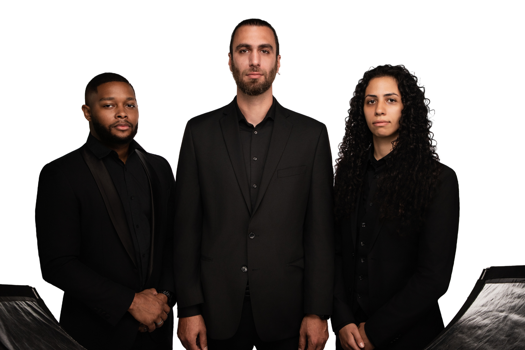 Three adults dressed in black suits standing against a white background, looking directly at the camera.