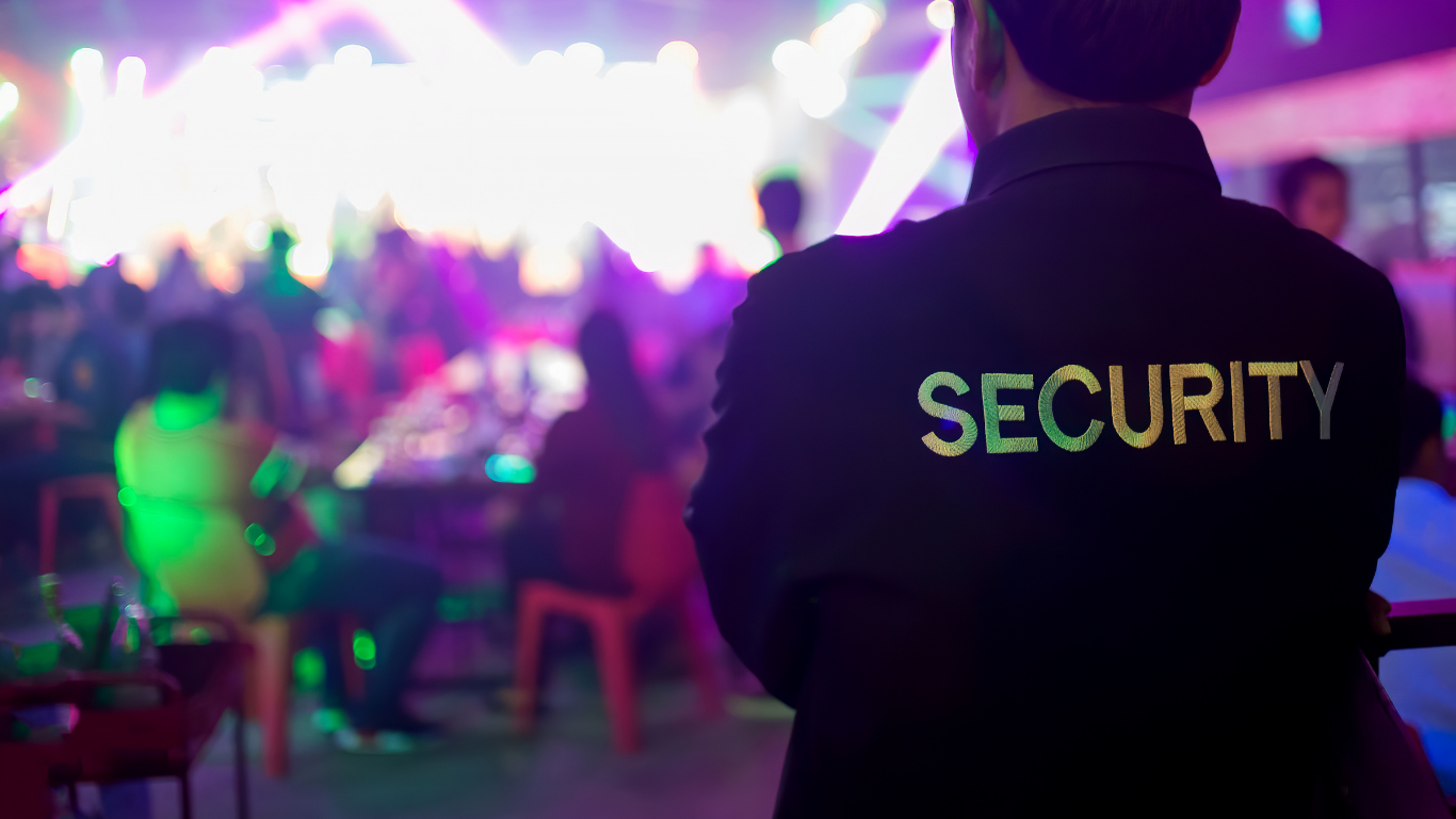 A security guard standing with arms crossed in a dark setting with colorful lights, at what appears to be a lively event or party with people sitting at tables and socializing.
