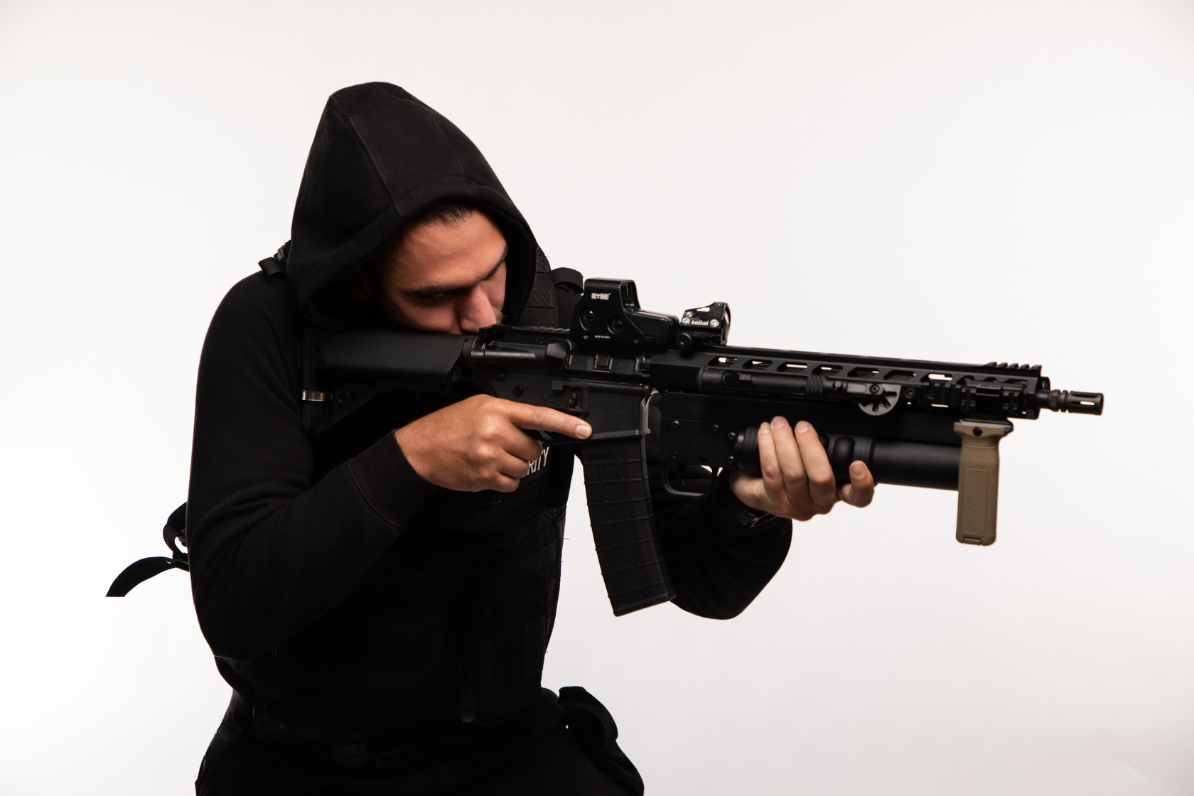 A person aiming a tactical rifle, wearing a black hoodie and tactical vest, against a plain white background.