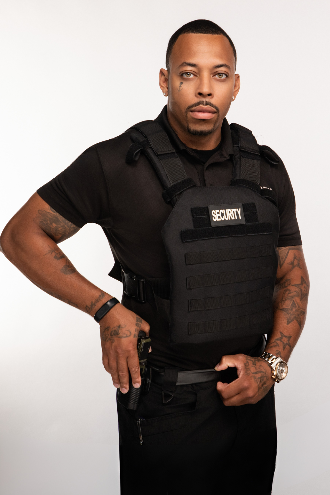 A man dressed as security personnel wearing a black uniform with a security vest, standing against a plain white background.