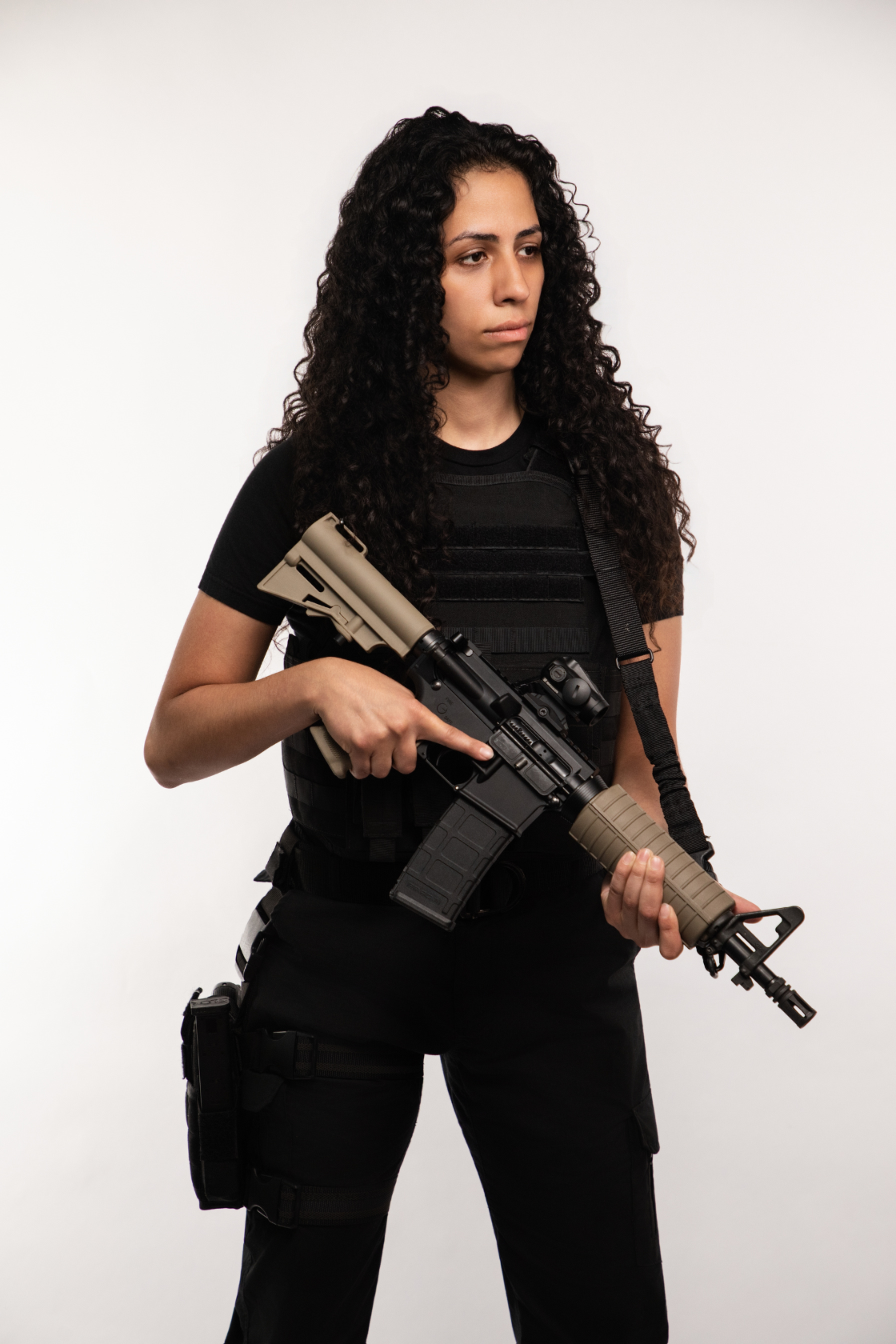 A woman dressed in black tactical gear holding a rifle with a scope against a plain white background.