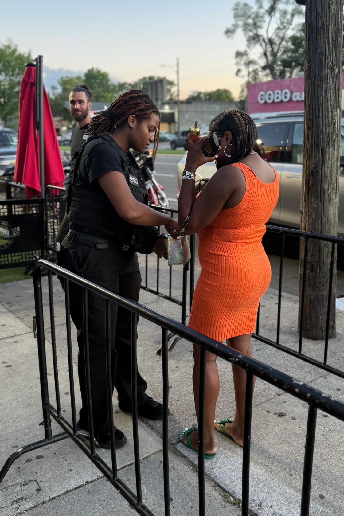 A woman in an orange dress is being searched by a female security officer outside a restaurant or store with a group of cars in the background.