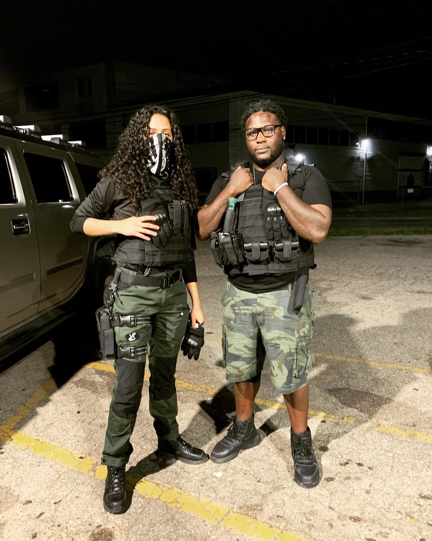 Two individuals in tactical gear standing outdoors at night, one woman and one man, with a vehicle in the background.