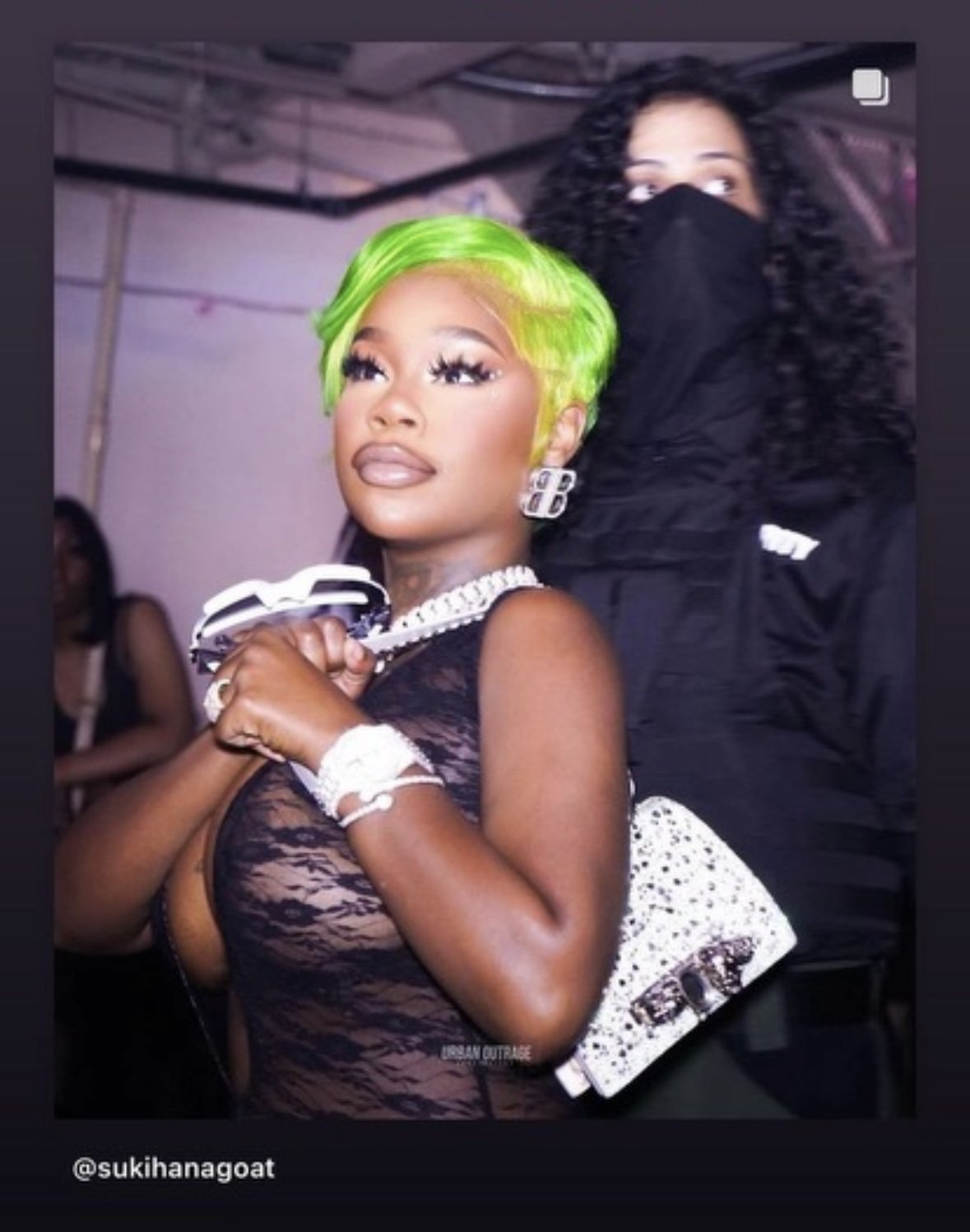 A woman with bright green hair, wearing a sheer black lace top, jewelry including a necklace, bracelet, large earrings, and holding sunglasses, poses at an event. She has makeup with defined eyes and glossy lips, and in the background, another woman 
