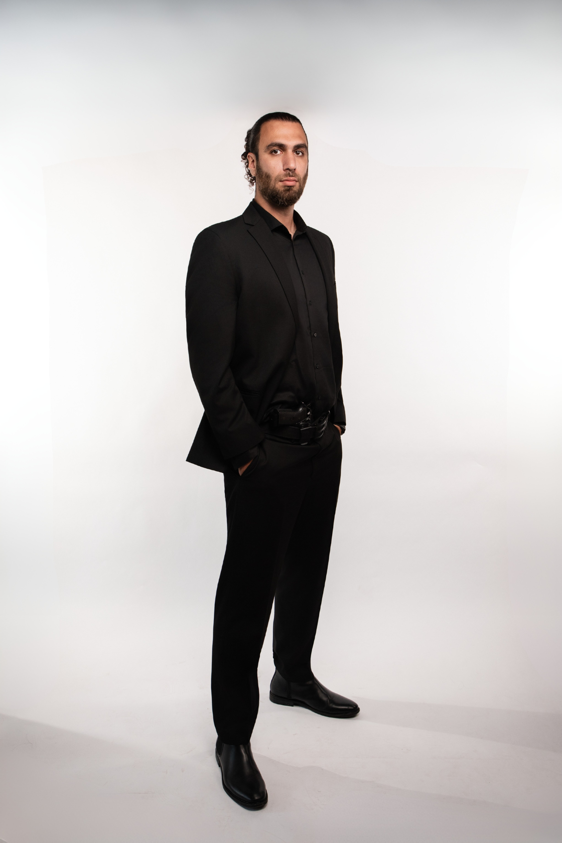 A man with dark hair and beard wearing a black suit, black shirt, and black shoes, standing with hands in pockets against a white background.