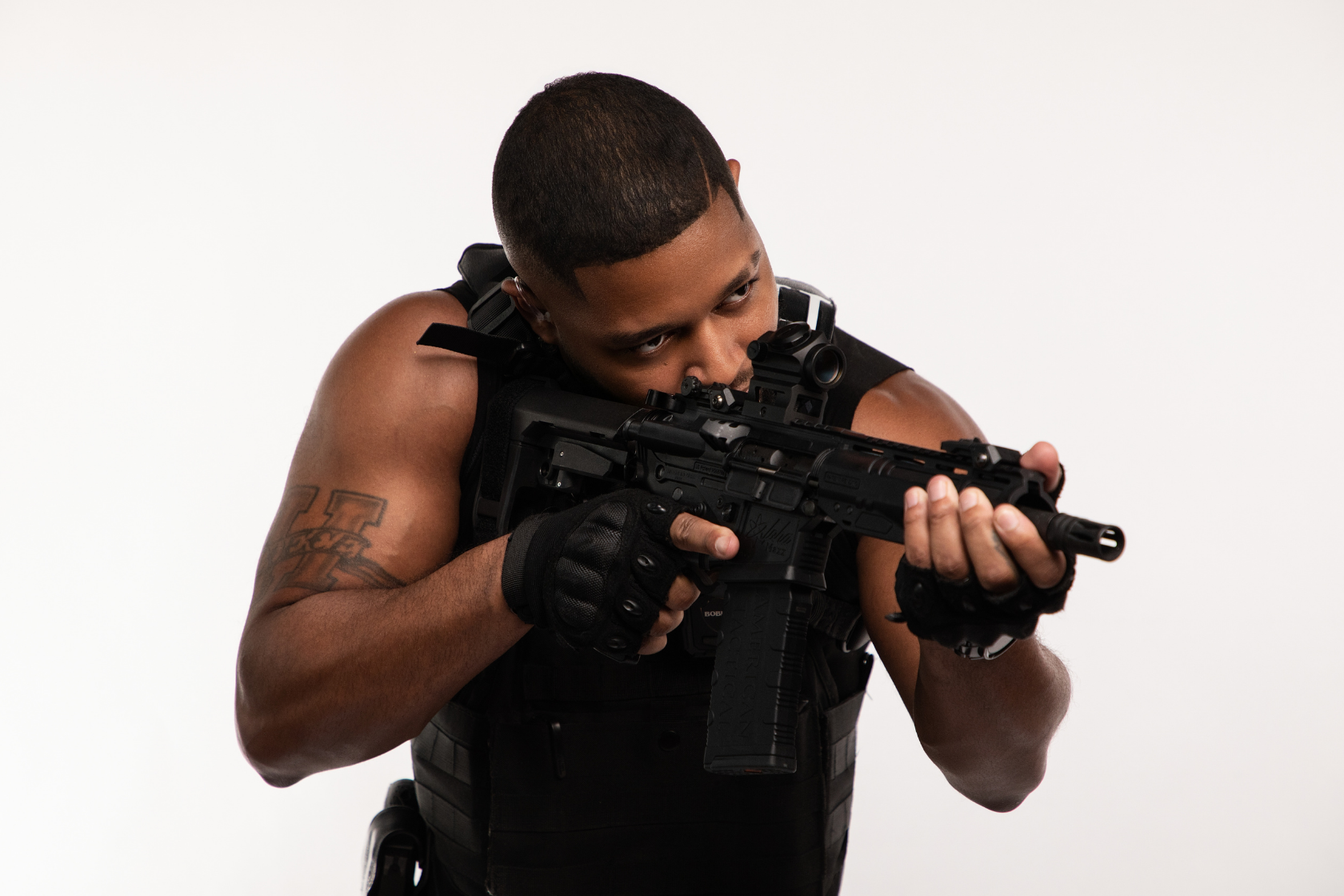 A man aiming a tactical rifle with a scope, dressed in black tactical gear, against a plain white background.