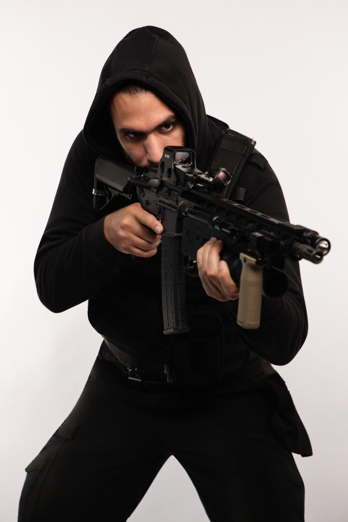 Person wearing black hoodie and tactical gear aiming a rifle.