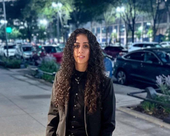 A young woman with long curly hair standing on a city sidewalk at night, with parked cars and trees in the background.