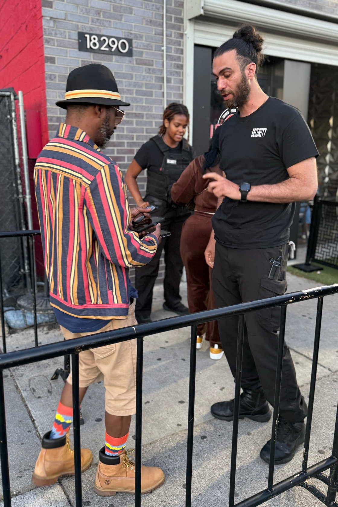 Two men are standing outside near a building, one dressed in a colorful striped shirt, tan shorts, and tan boots, looking at his phone. The other man, wearing a black security uniform and black pants, is talking to him. A woman dressed in black is st