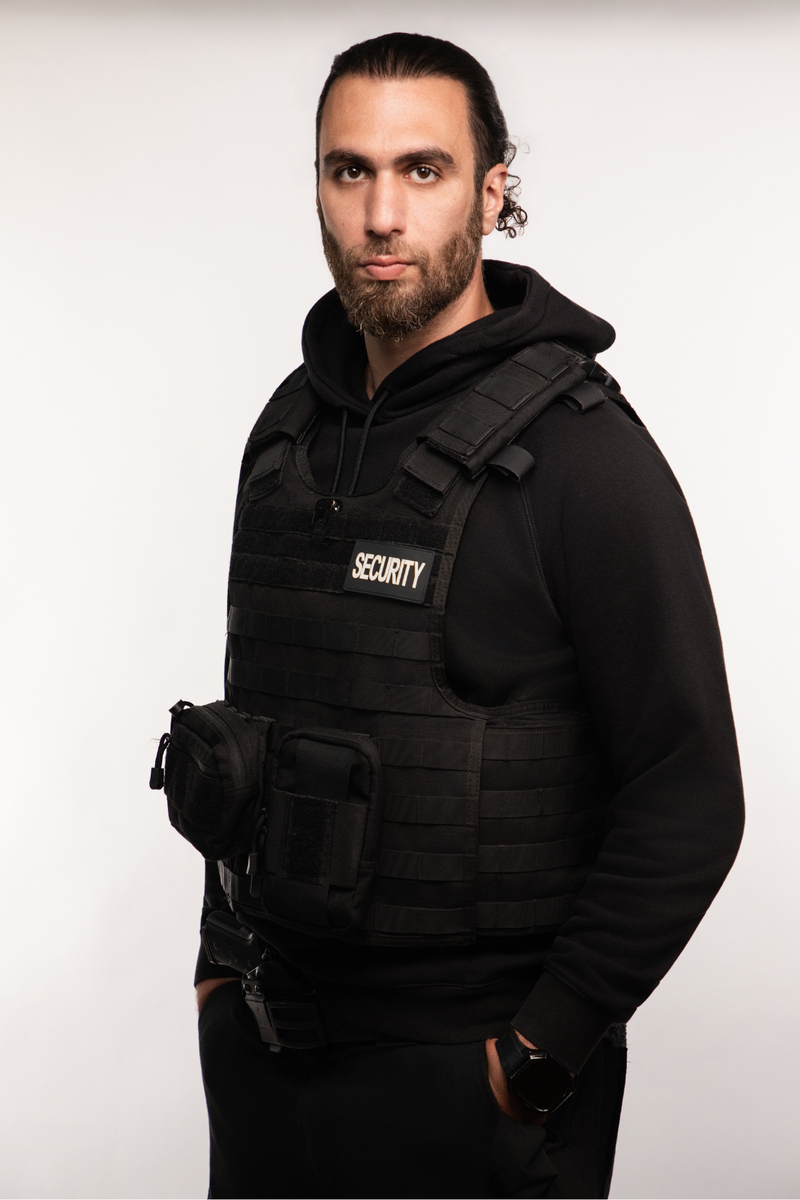 Man in black security tactical vest with hands in pockets standing against a plain white background.