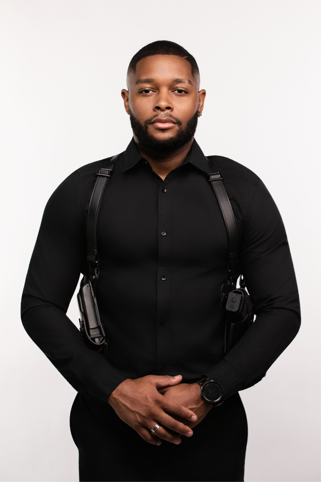 A young man with a beard and short hair wearing a black shirt and a wristwatch, standing against a white background.