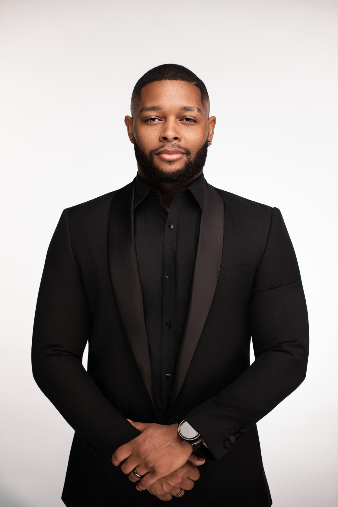 A man wearing a black suit with a black shirt and a watch, standing against a plain white background.