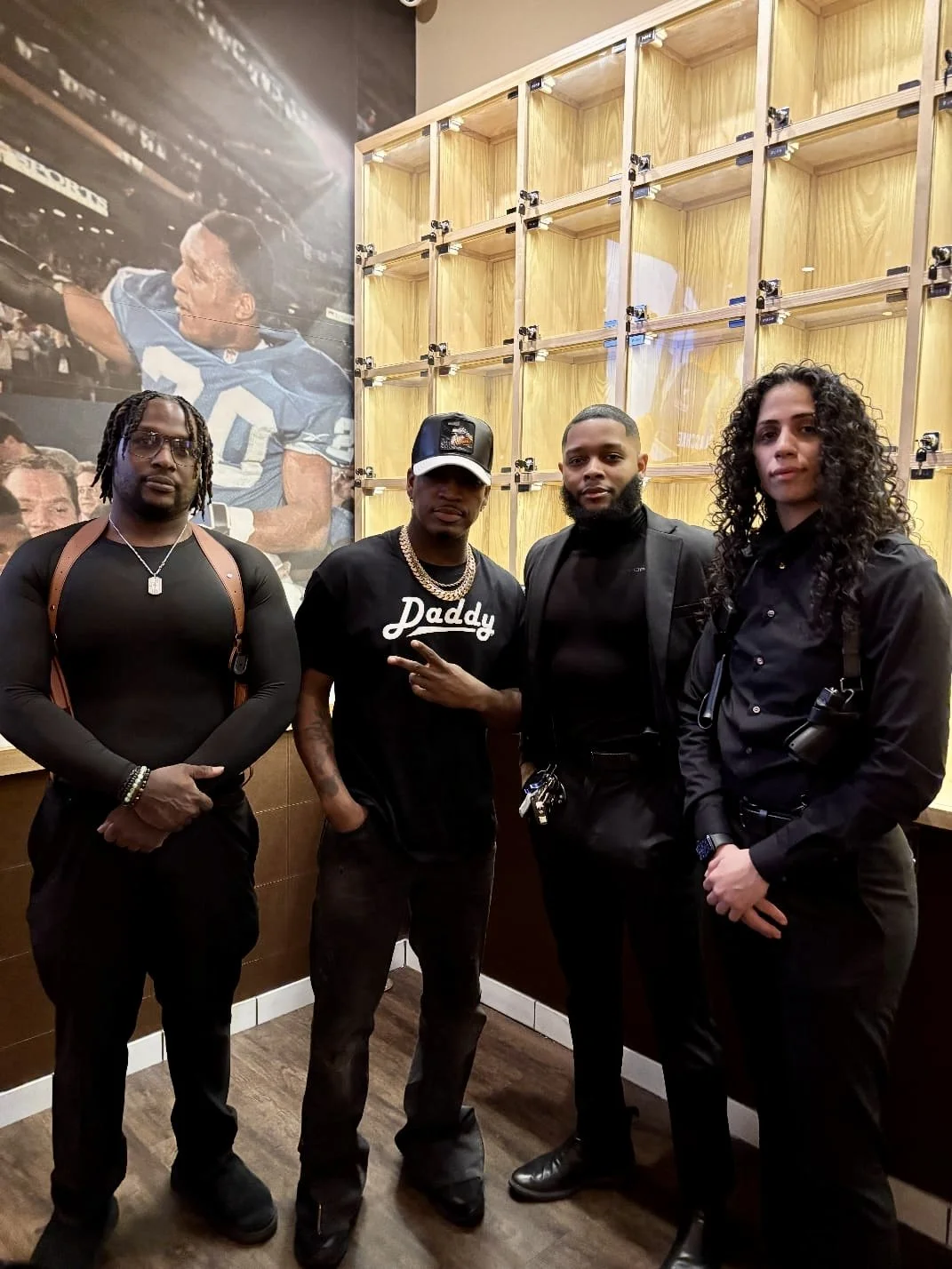 Four people standing indoors in front of a wooden shelf and a large poster of a football player.