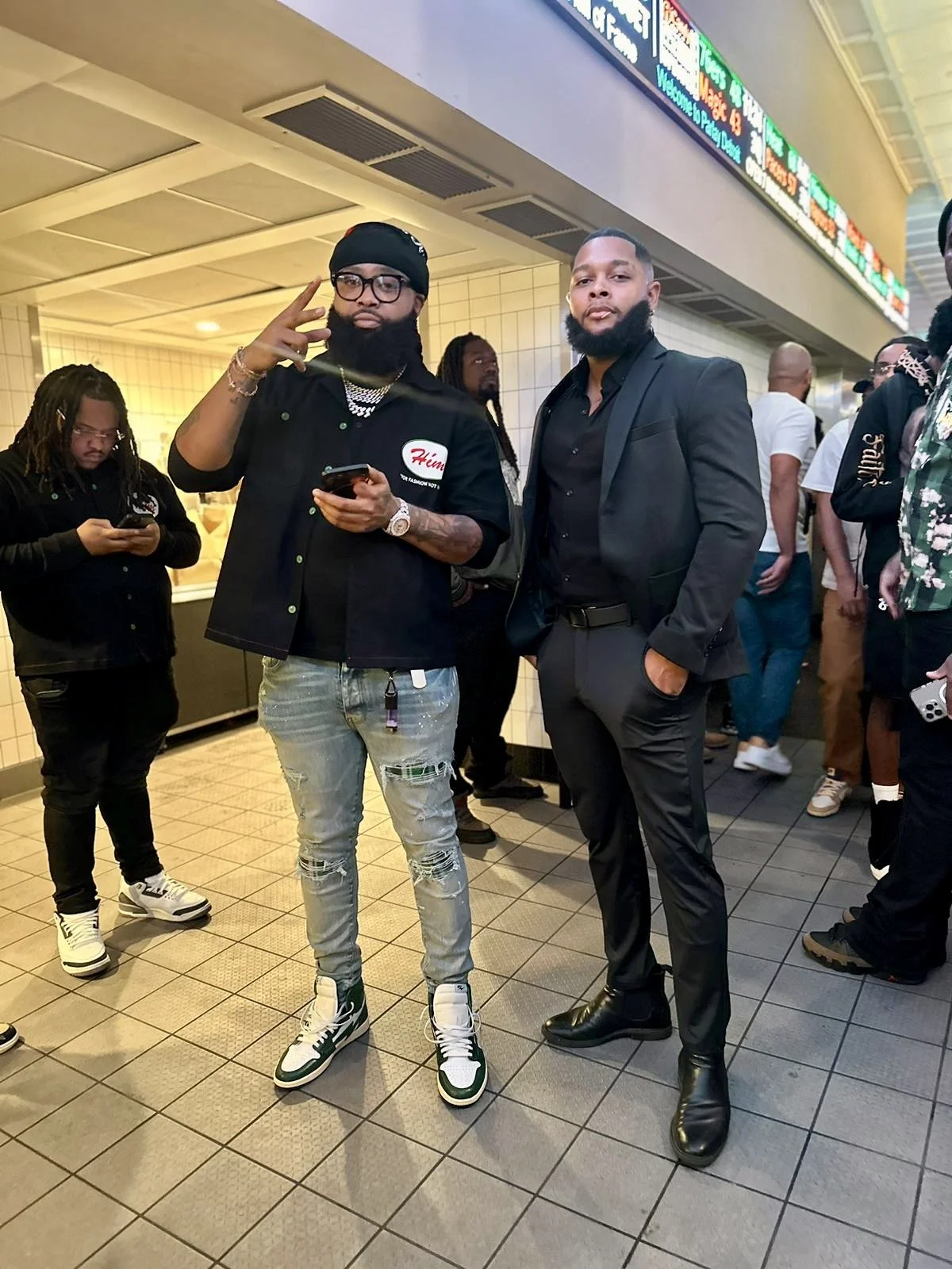 Two men in fashionable attire posing inside a busy public space, likely an airport or train station, with other people in the background, some using smartphones.