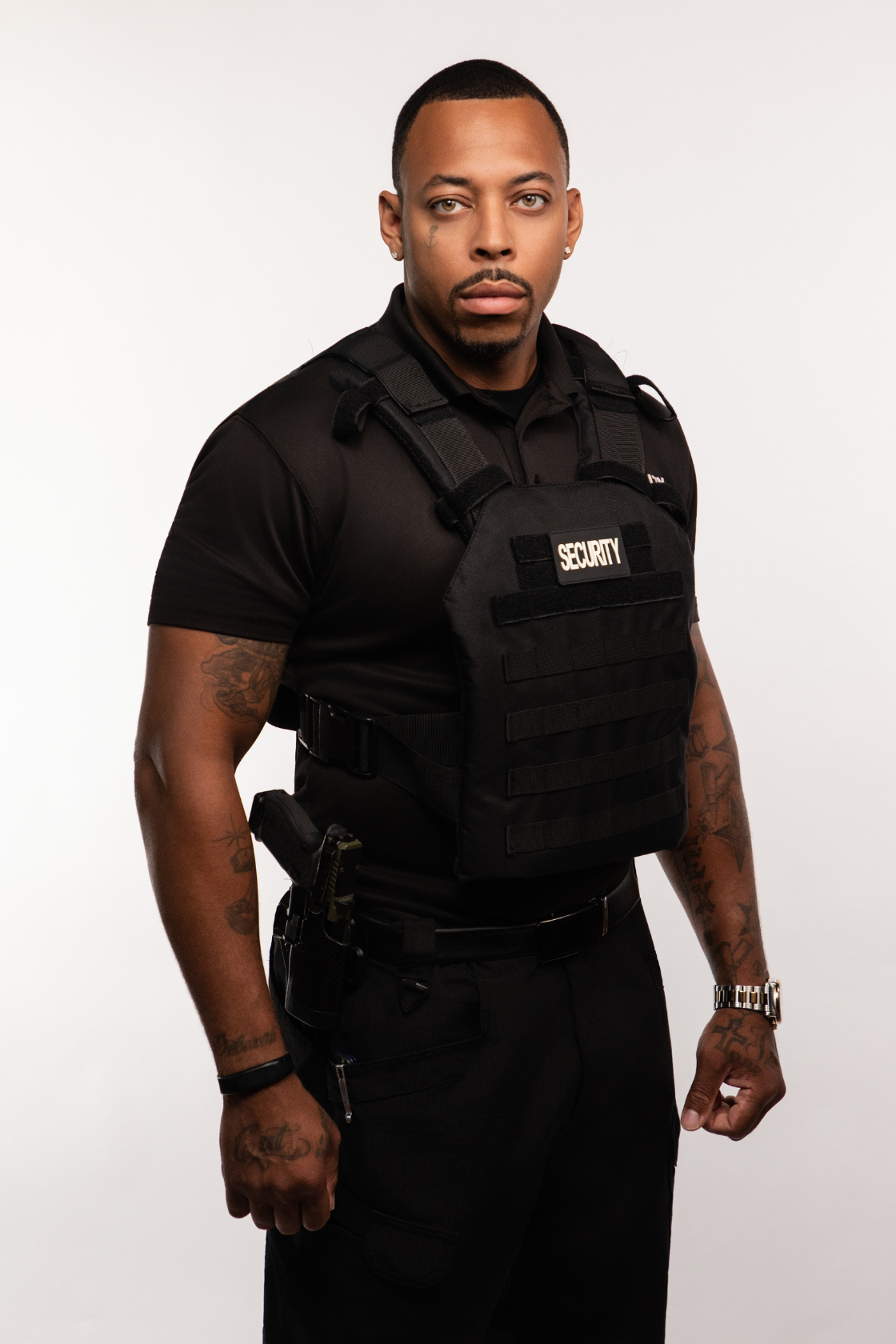 A male security guard wearing a black uniform, vest labeled 'SECURITY', with a gun holstered on his waist, standing against a plain white background.