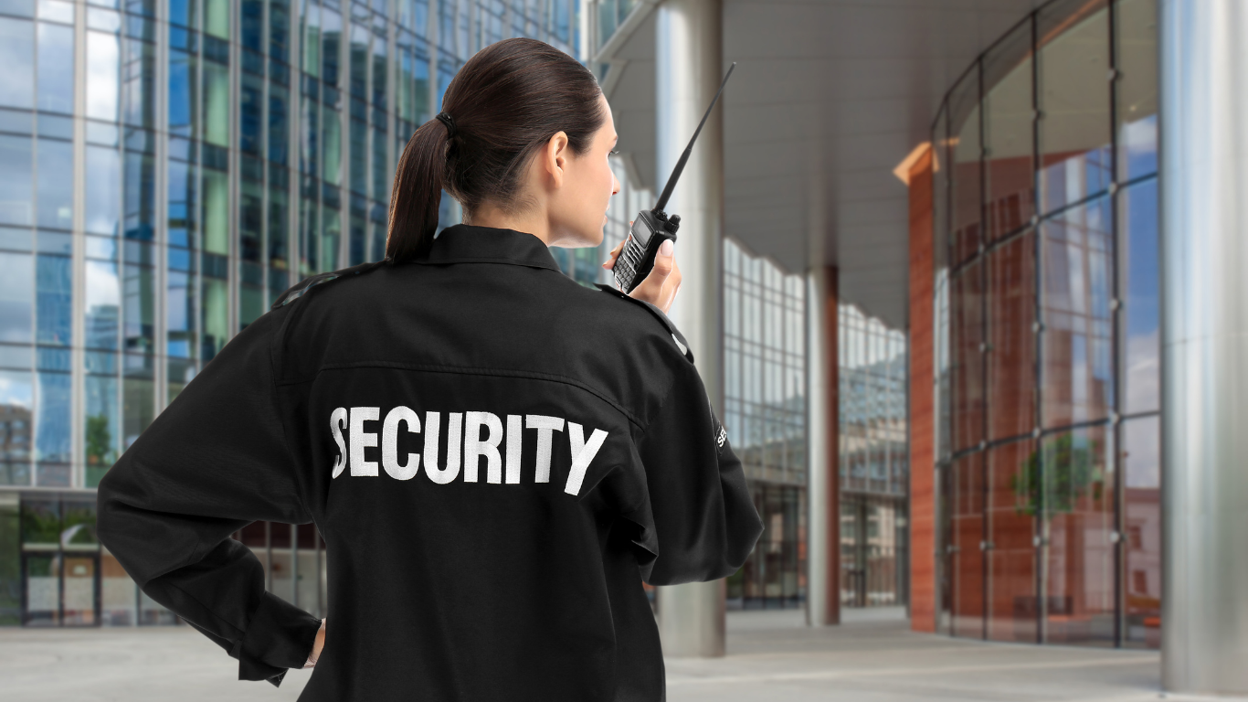 Security guard in black uniform with 'SECURITY' label, holding walkie-talkie outside modern glass building.
