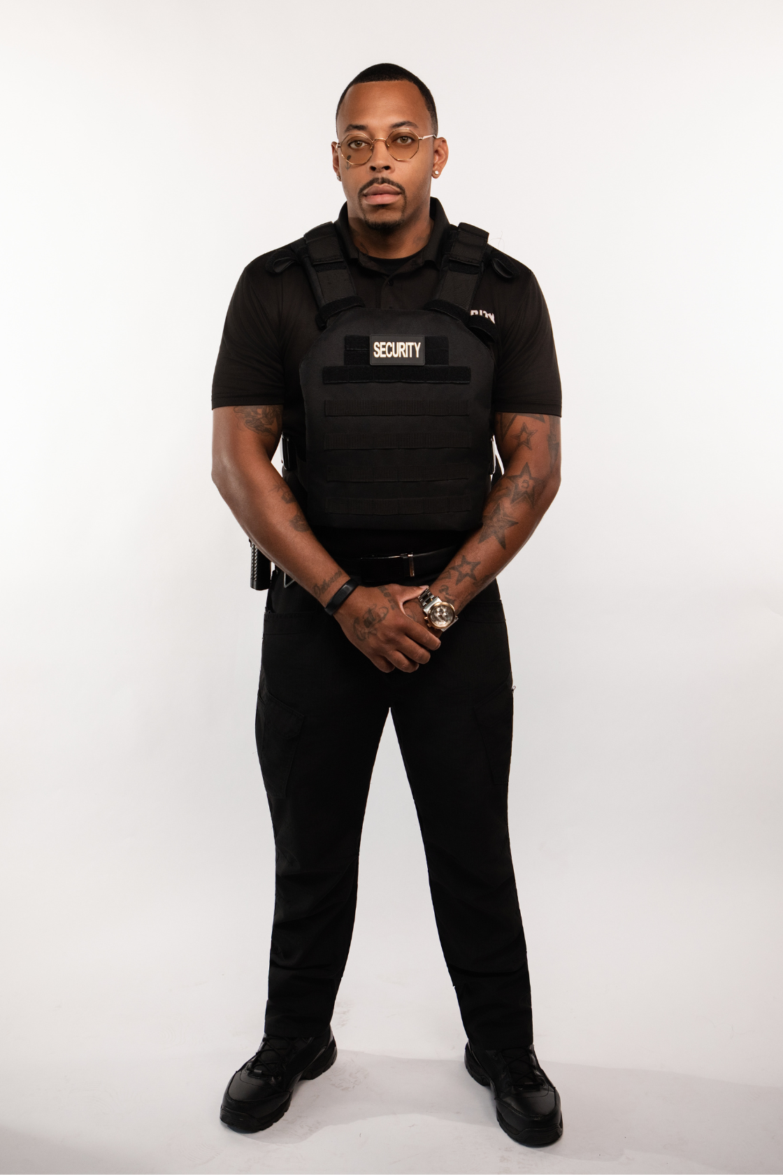 A security guard wearing glasses, a black polo shirt, a tactical vest with the word 'SECURITY' on it, and black pants, standing with hands clasped in front against a plain white background.