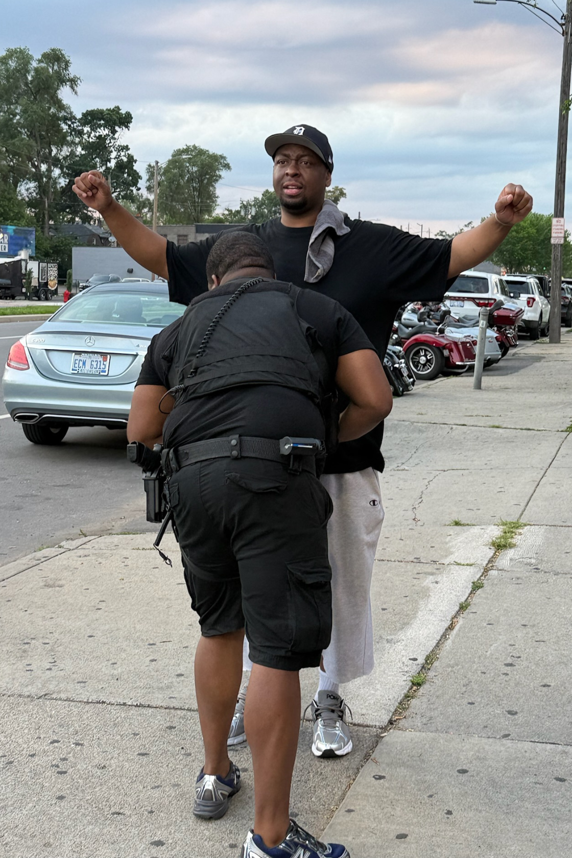 A man with a gray towel around his neck is being detained by a security or police officer outdoors on a sidewalk, with parked cars and motorcycles in the background under a cloudy sky.