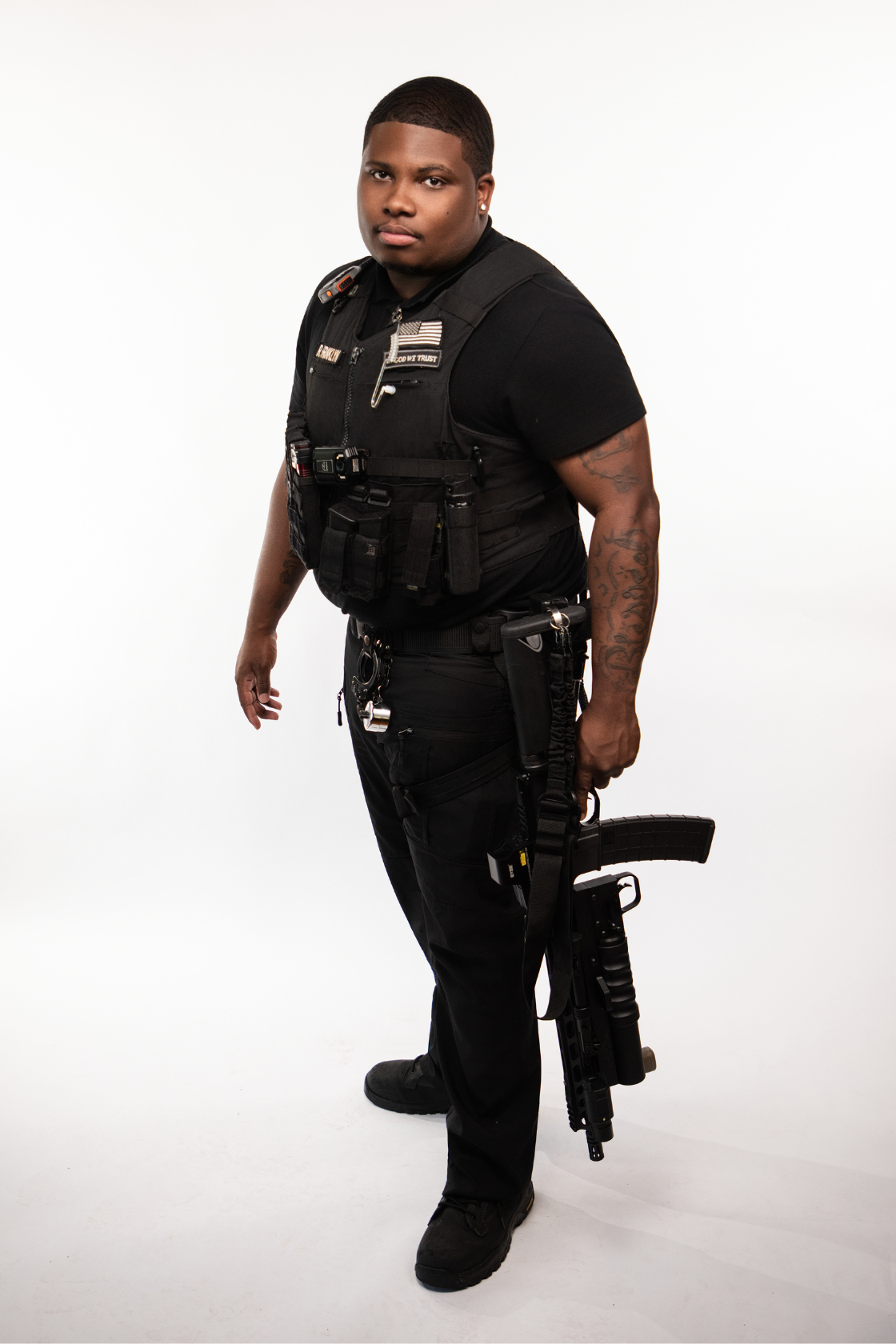 A young African American man dressed in black tactical gear, holding a large firearm, standing against a plain white background.