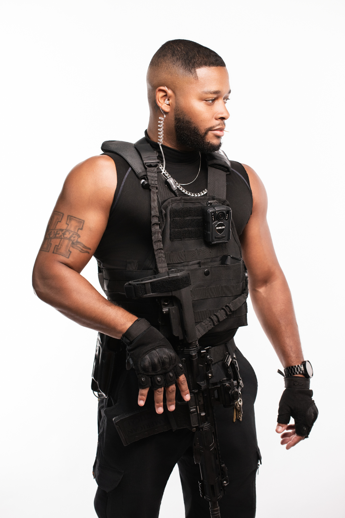 A man dressed in tactical gear, including a bulletproof vest, gloves, and carrying a firearm, with a headset and a radio attached to his vest.