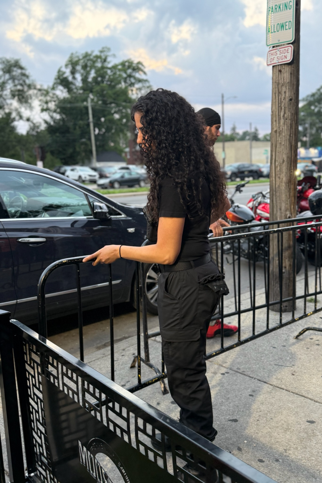 A woman with curly dark hair wearing a black t-shirt and cargo pants standing next to a black shopping cart on a sidewalk, with parked cars and motorcycles nearby and a wooden pole with parking signs in the background.