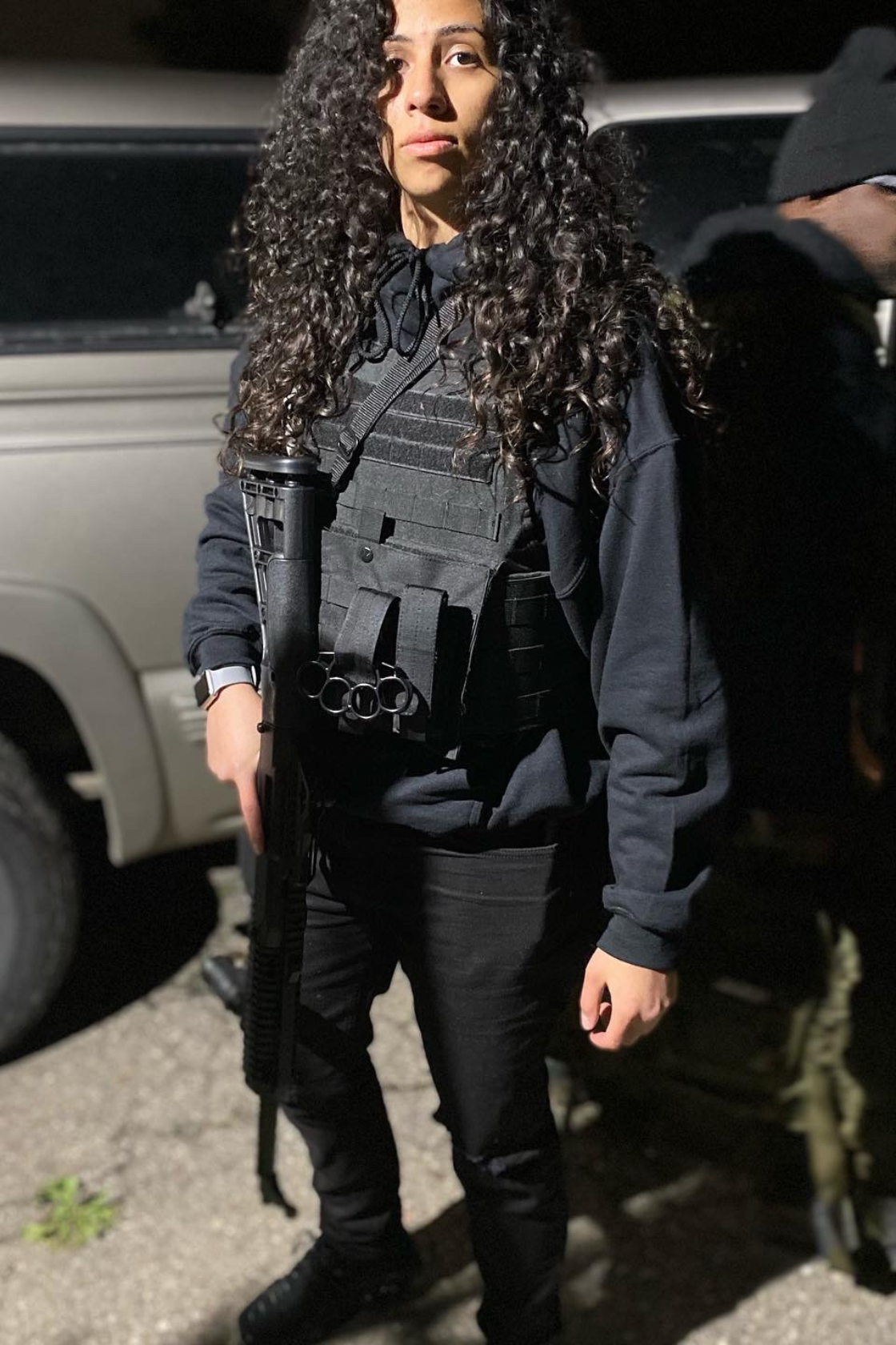 A woman with long, curly dark hair stands outdoors at night, dressed in black tactical gear, holding a firearm, with a serious expression.
